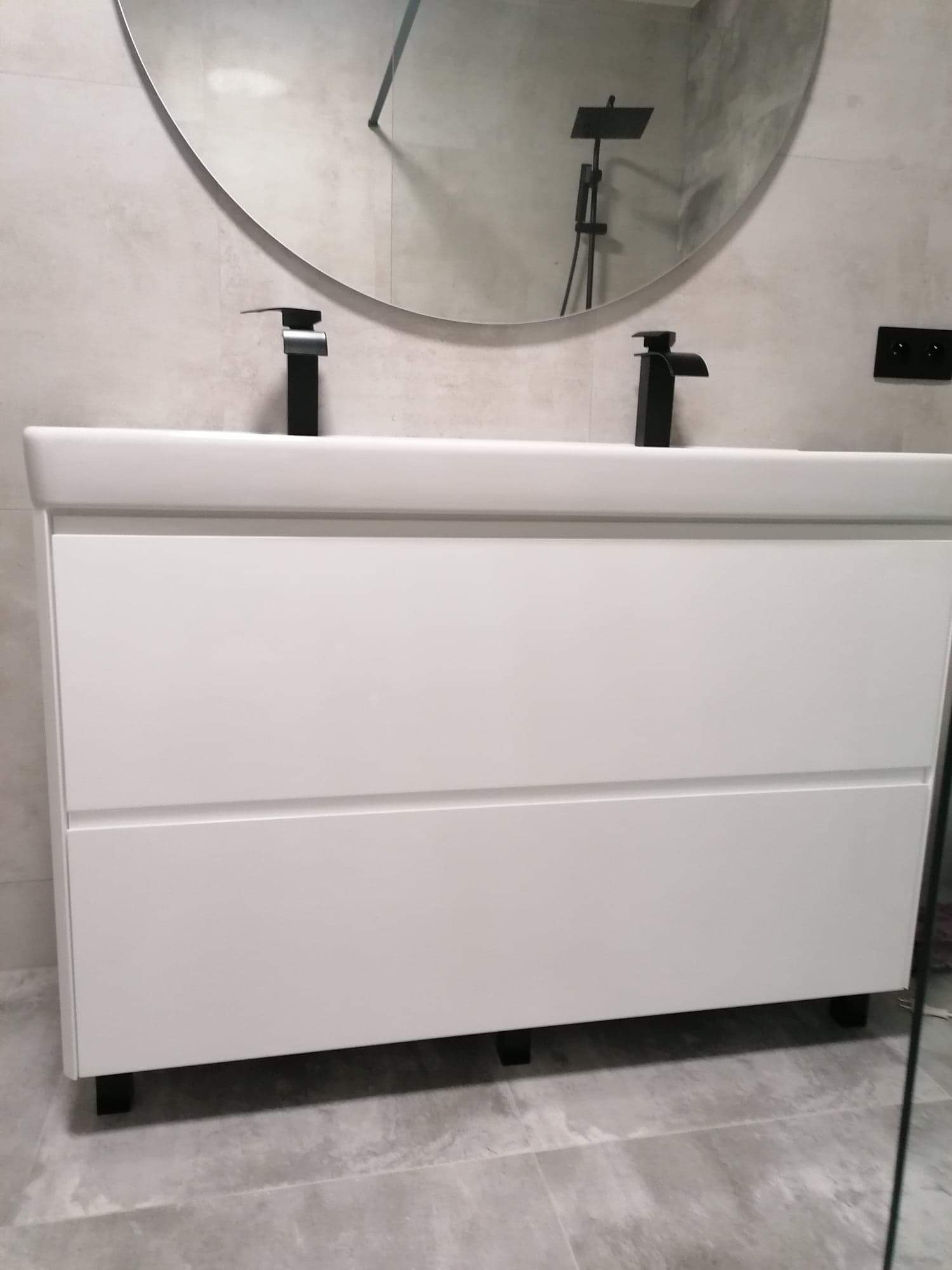 Biała szafka łazienkowa z dwiema szufladami i czarnymi bateriami, okrągłe lustro z odbiciem czarnej deszczownicy, szare płytki podłogowe i ścienne.