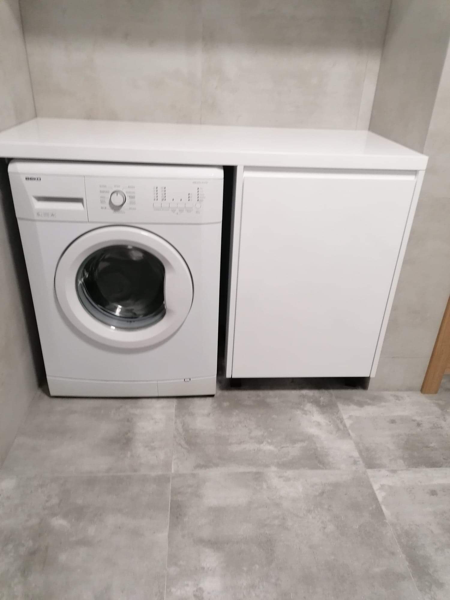 Pralka zabudowana w szafce na wymiar z blatem, szara płytka podłogowa, minimalistyczne wnętrze łazienki.