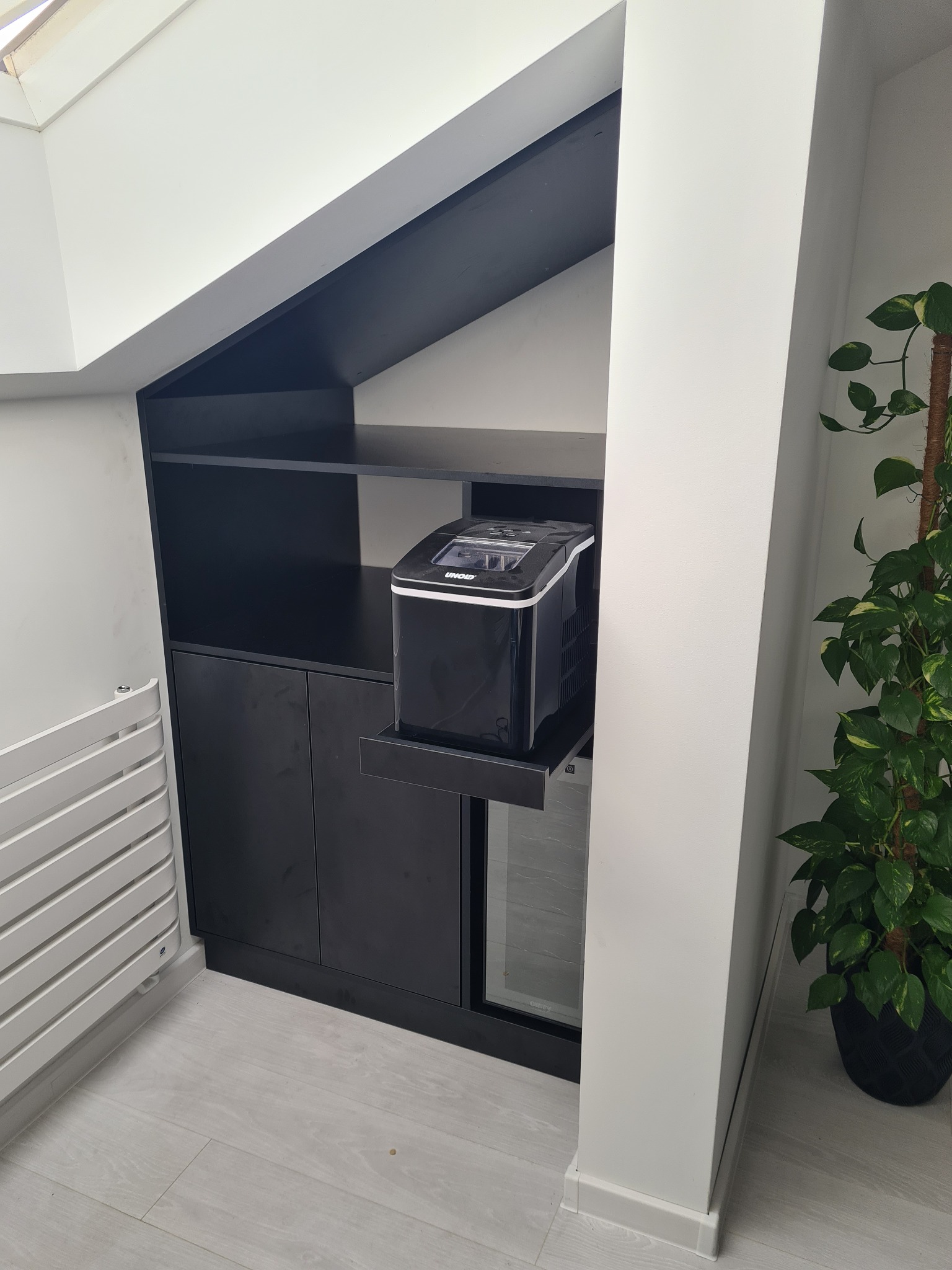 Czarna zabudowa meblowa pod skosem dachu z wysuwaną półką, na której stoi kostkarka do lodu, obok grzejnik i roślina doniczkowa.