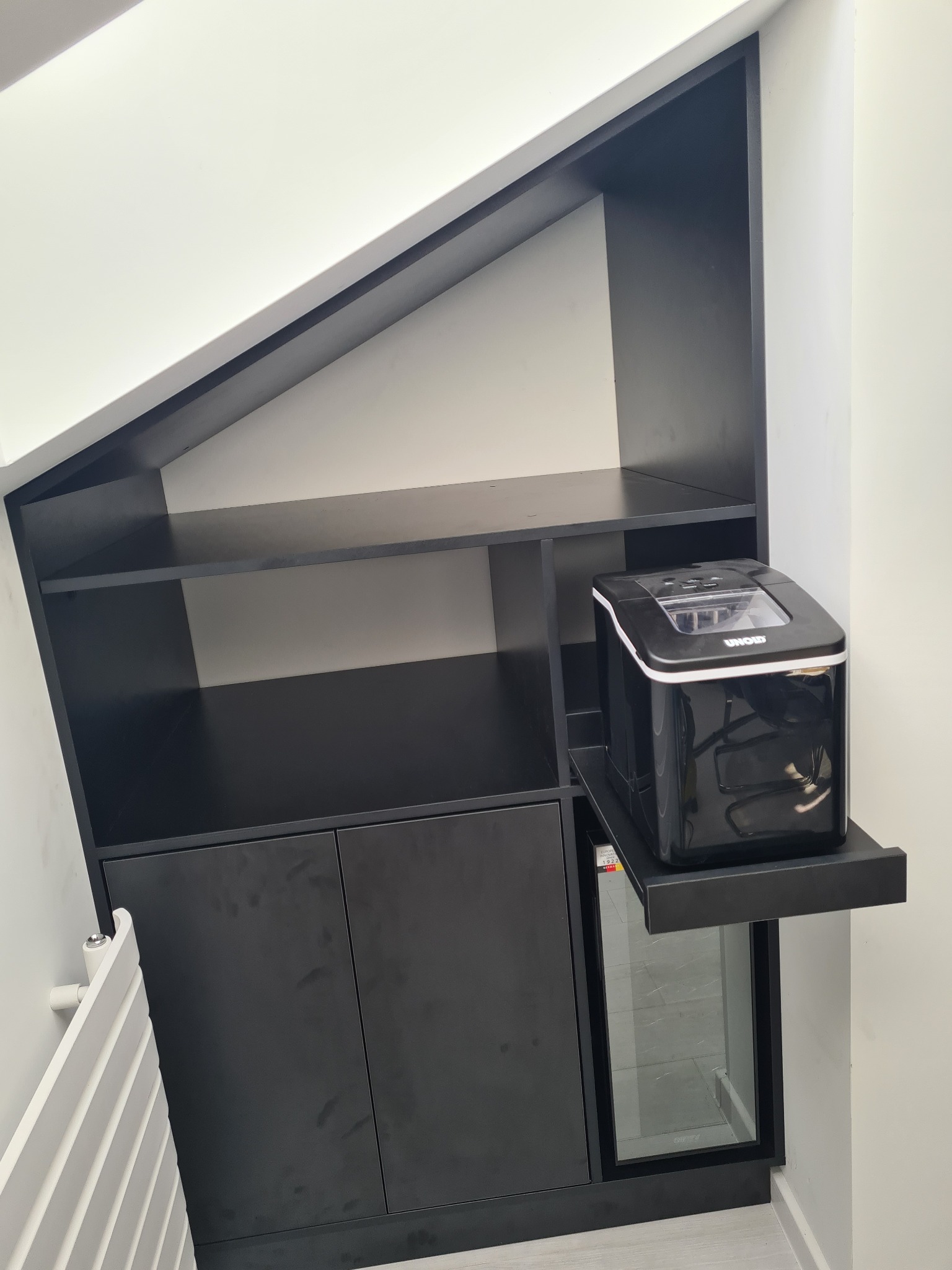 Czarna szafa na wymiar z półkami i szafkami, wbudowana w skos dachu, z dodatkową wysuwaną półką na kostkarkę do lodu.