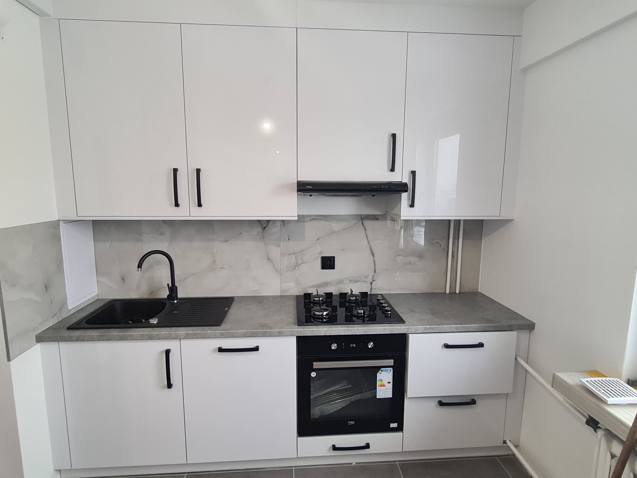 Biała, nowoczesna zabudowa kuchenna z błyszczącymi frontami, czarnymi uchwytami i blatem imitującym beton, uzupełniona o czarny zlew, płytę grzewczą i piekarnik.