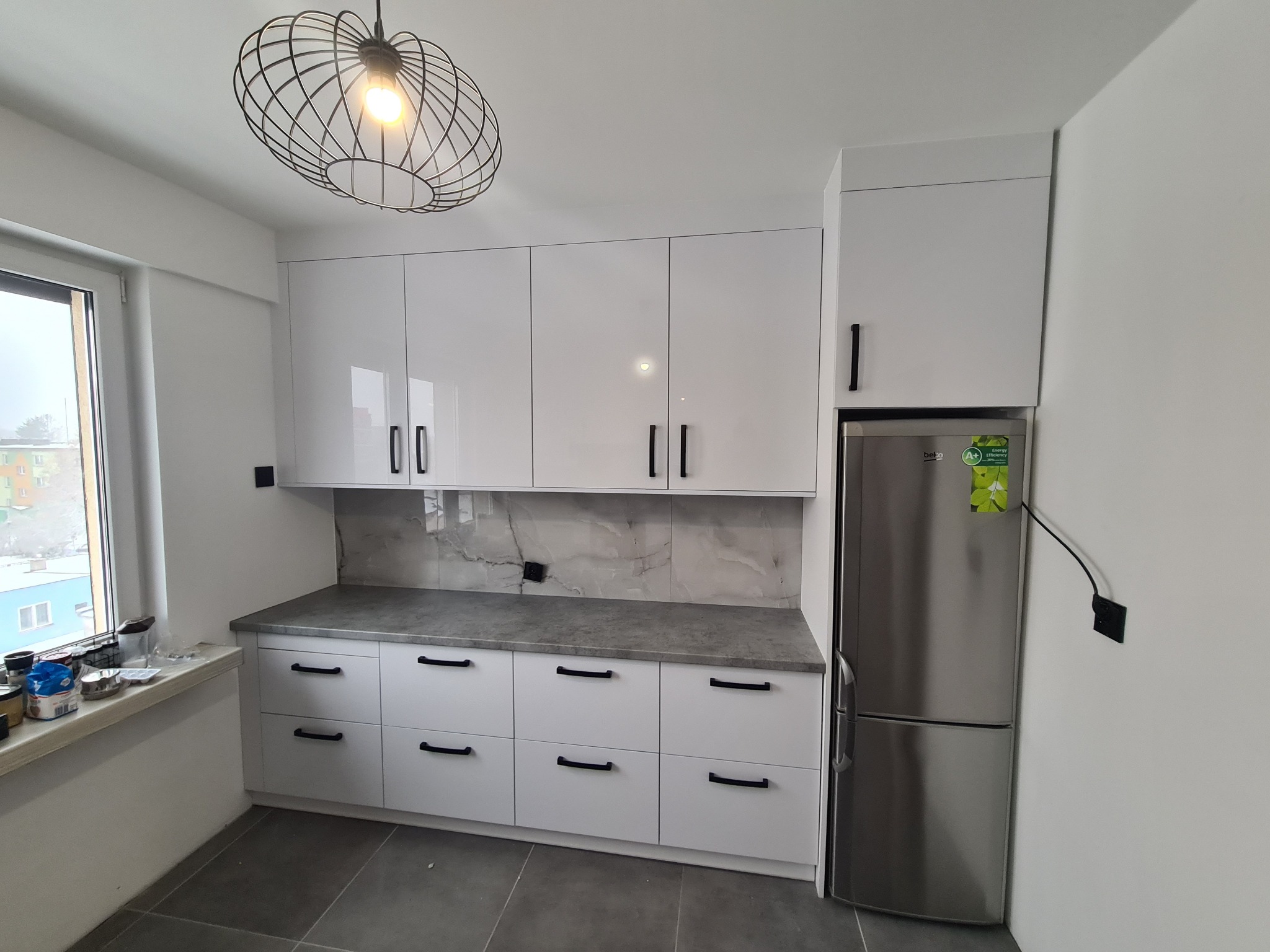 Nowoczesna kuchnia z białymi, lakierowanymi szafkami z czarnymi uchwytami, szarym blatem i marmurowym backsplashiem, widoczny fragment okna z akcesoriami kuchennymi.