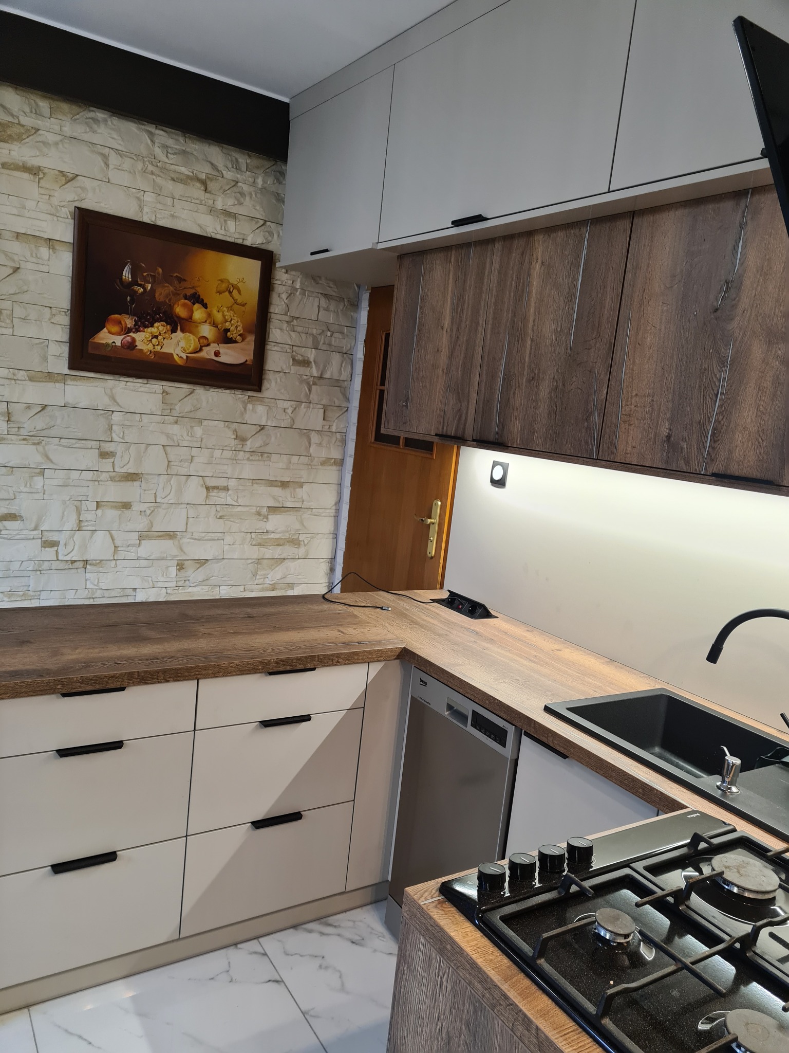 Nowoczesna kuchnia z białymi szafkami dolnymi i górnymi w kolorze drewna, czarnymi uchwytami, blatem z drewnopodobnego laminatu, płytą gazową i zlewozmywakiem, na ścianie dekoracyjna cegła i obraz...