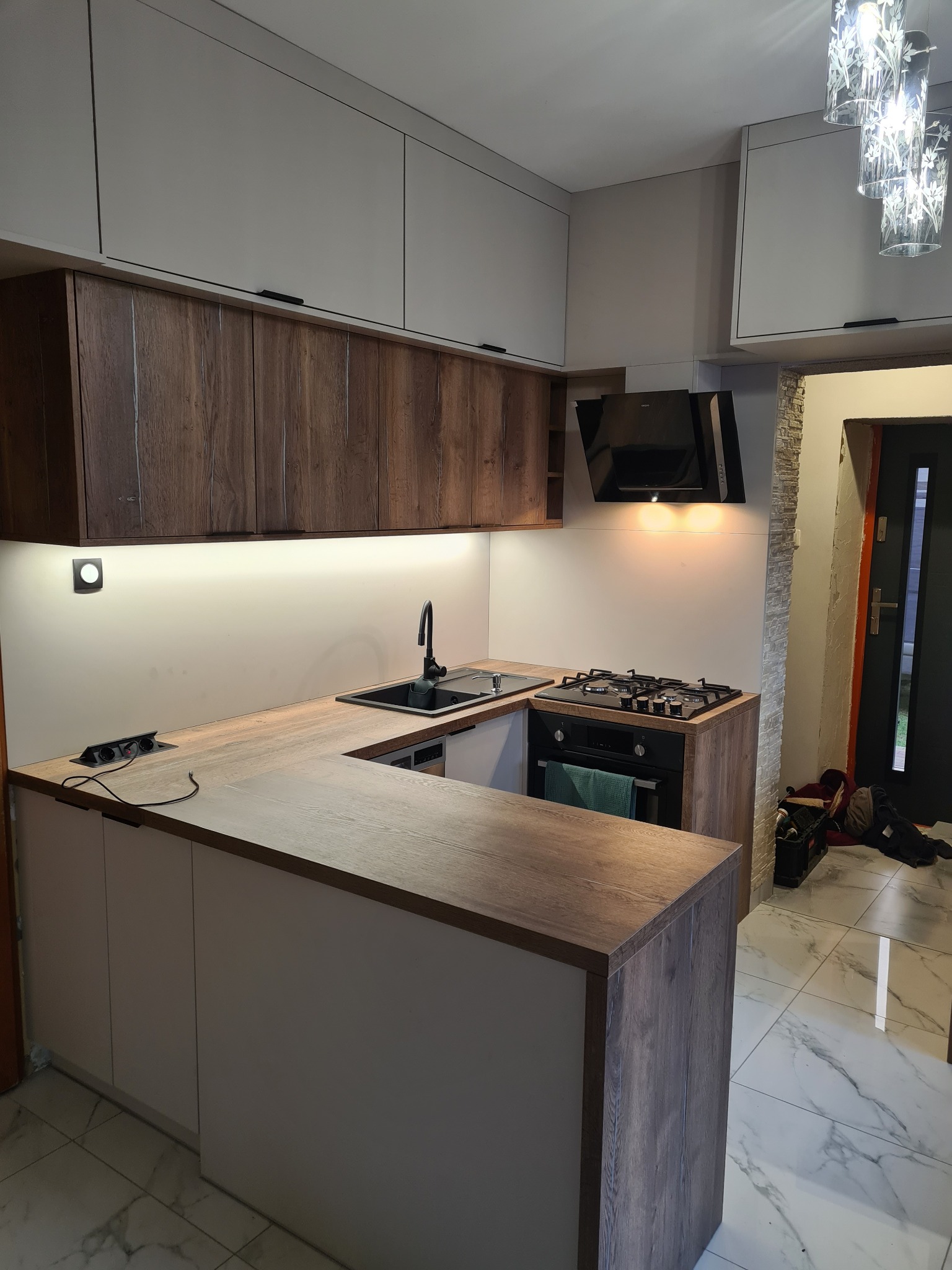 Nowoczesna kuchnia w odcieniach bieli i drewna z czarnymi akcentami, oświetleniem LED pod szafkami wiszącymi i marmurową podłogą.