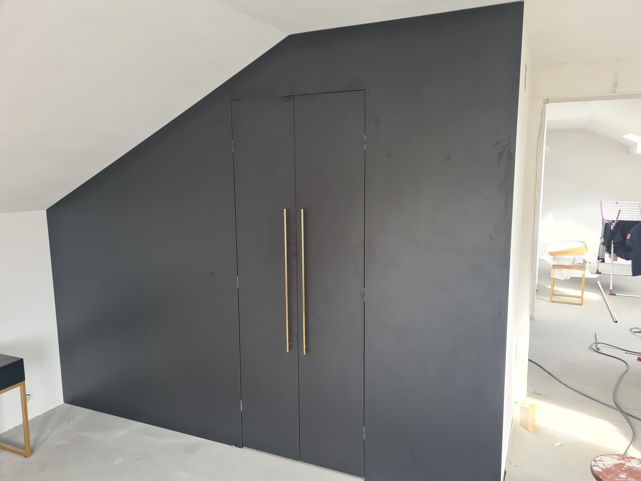 Grafitowa szafa wnękowa z długimi, złotymi uchwytami, wkomponowana w skos dachu, widok z boku na surową przestrzeń w trakcie wykończenia.