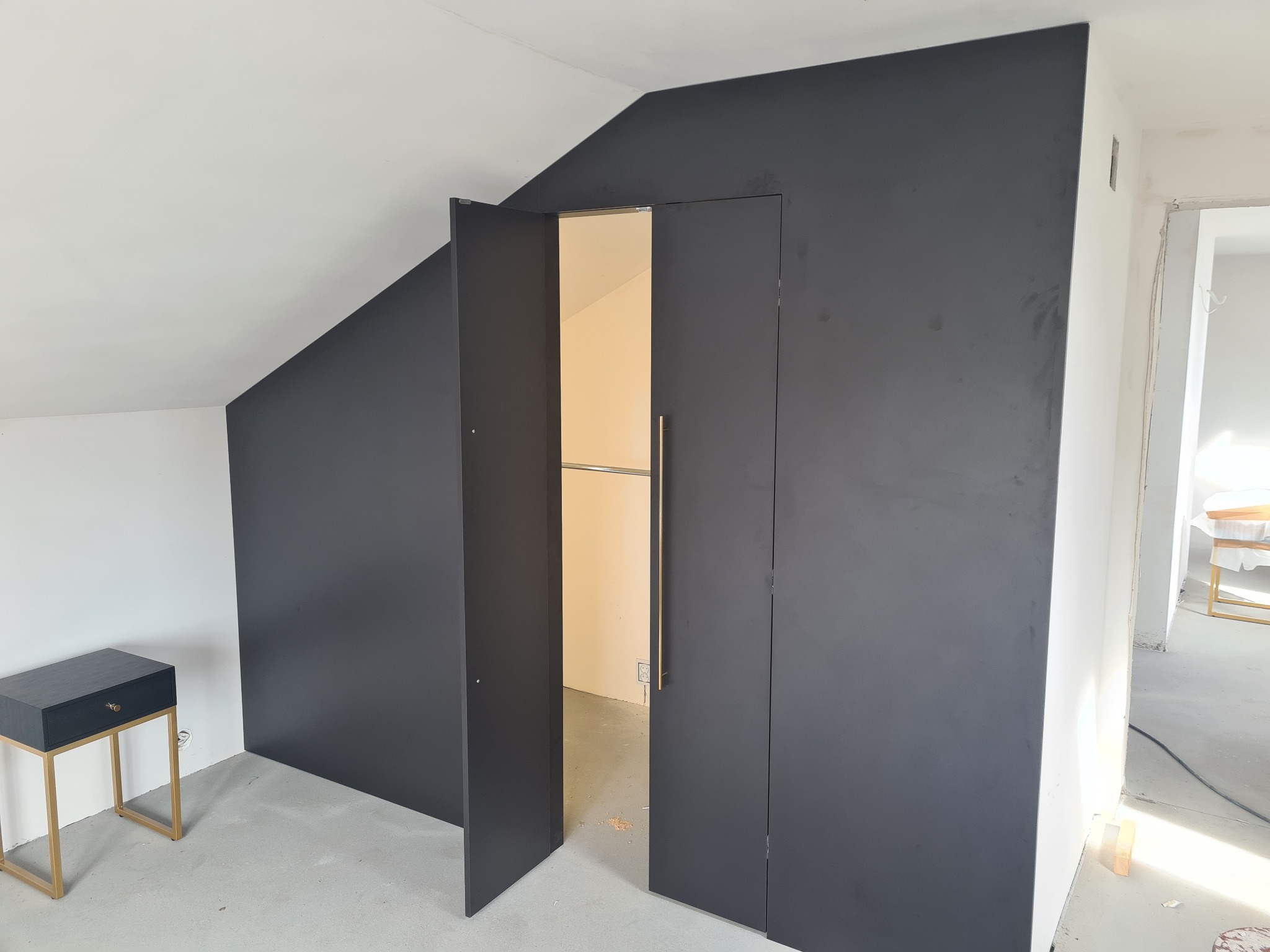 Czarna szafa wnękowa z otwartymi drzwiami w skosie poddasza, widoczny drążek na ubrania i mała szafka nocna w podobnym stylu.