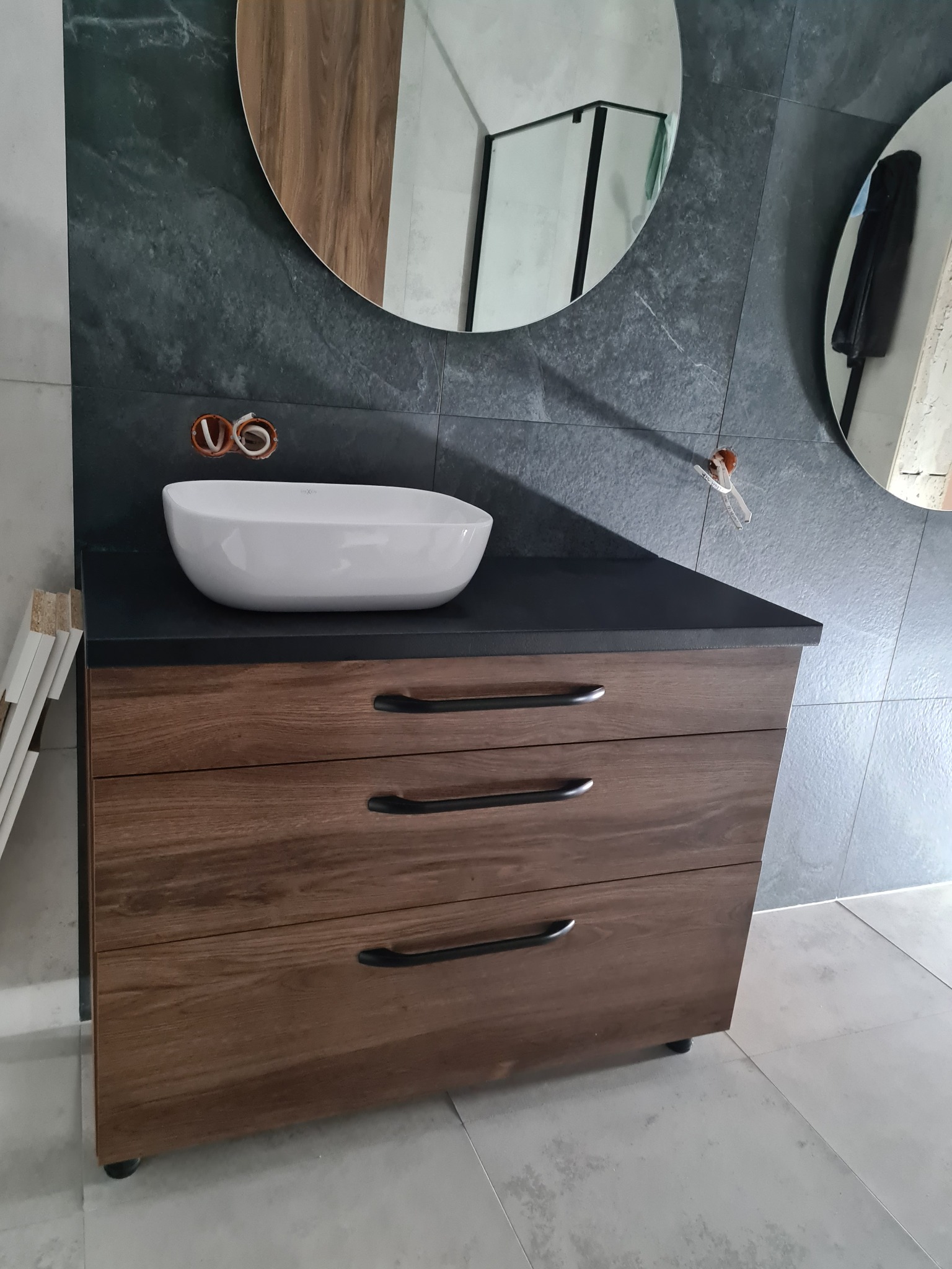 Łazienkowa szafka z trzema szufladami o drewnianym wykończeniu, z czarnym blatem i białą umywalką nablatową. W tle szara ściana z dwoma okrągłymi lustrami i widocznymi instalacjami elektrycznymi...