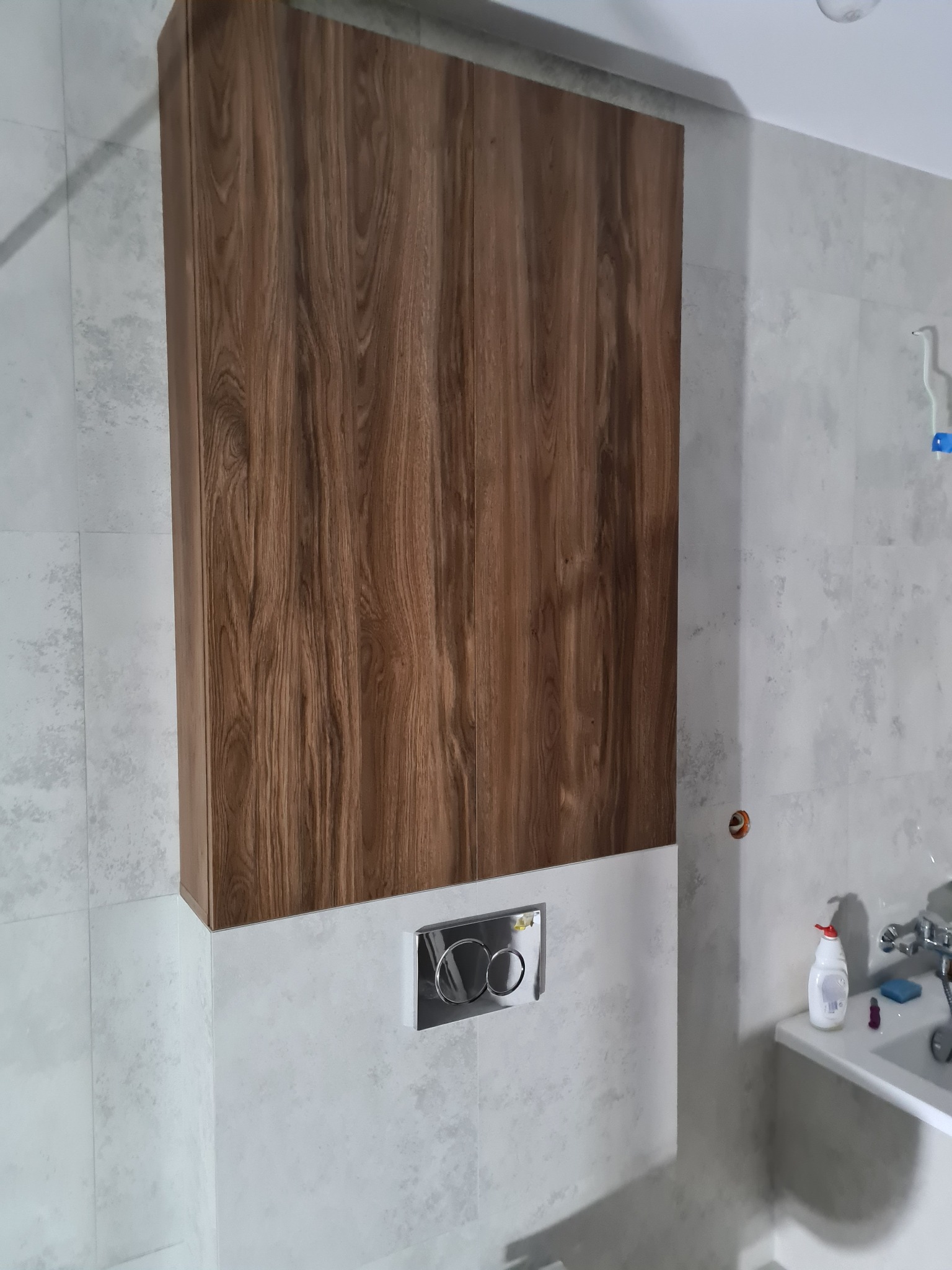 Wysoka szafka łazienkowa z drewnianym frontem, zamontowana nad spłuczką w toalecie wykończonej jasnymi, wzorzystymi płytkami.