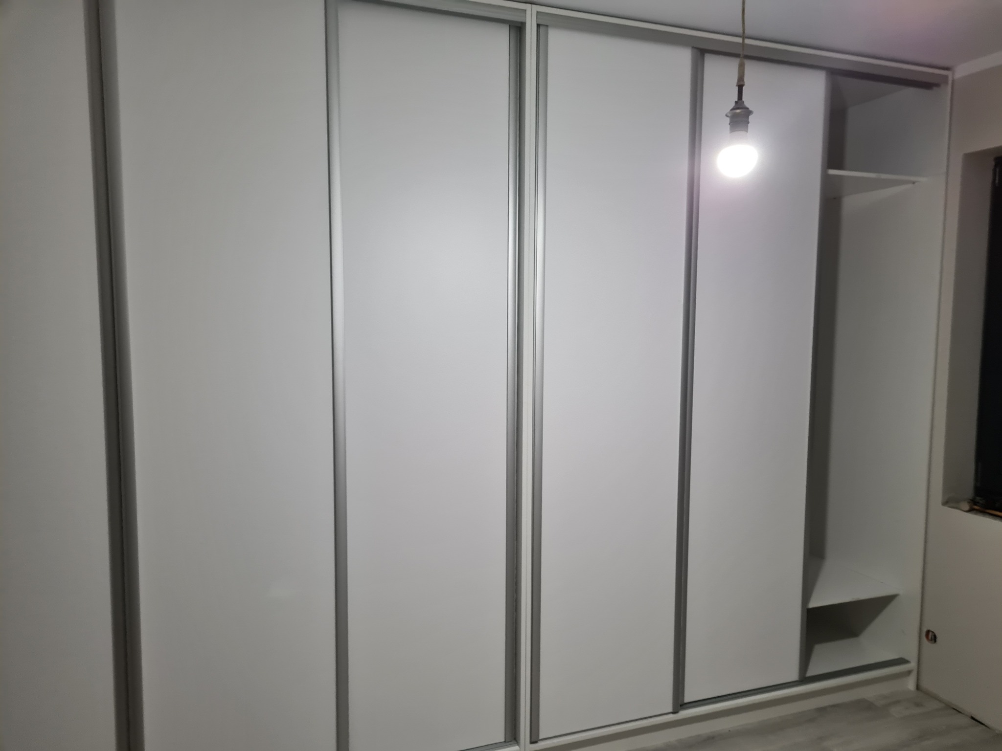 Biała szafa wnękowa z przesuwnymi drzwiami i widocznymi półkami z boku, oświetlona wiszącą żarówką na kablu.