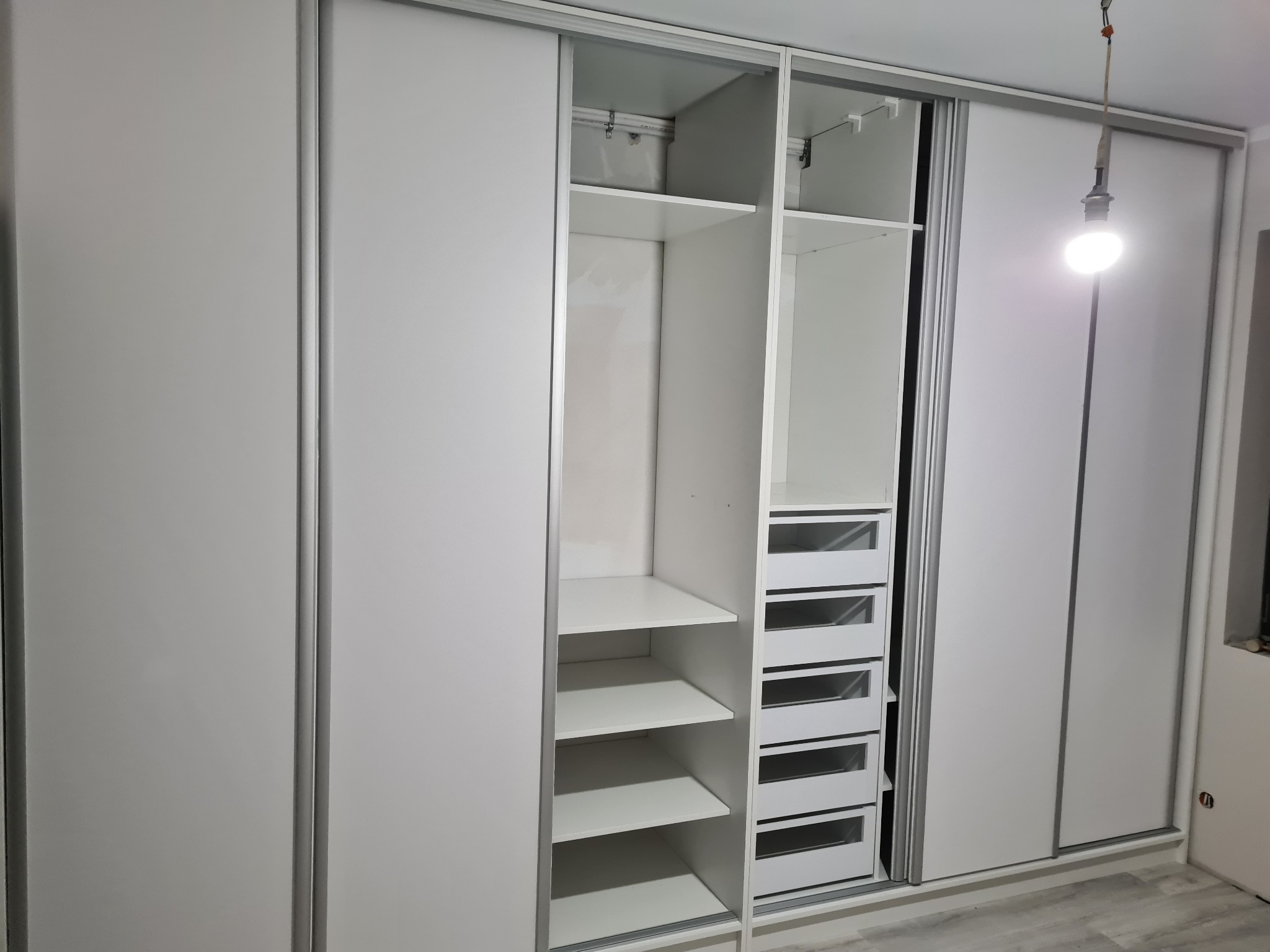 Biała szafa wnękowa z przesuwnymi drzwiami, częściowo otwarta ukazująca półki i szuflady, oświetlona wiszącą żarówką.