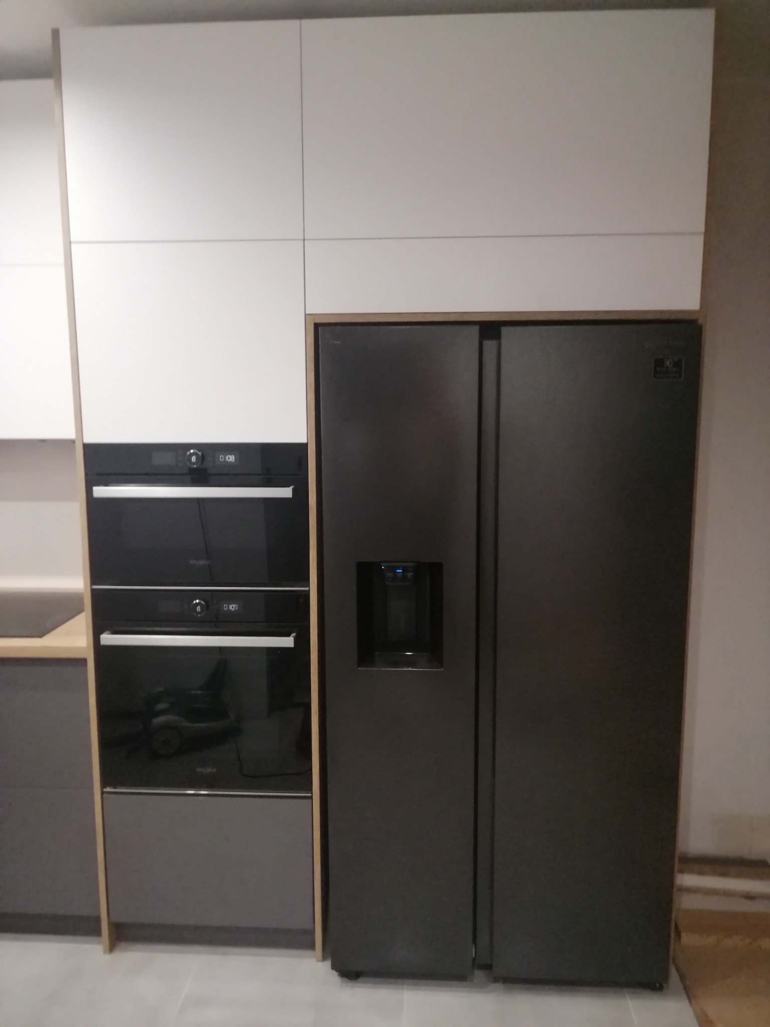 Nowoczesna zabudowa kuchenna z białymi szafkami, piekarnikami w czarnej obudowie i dużą, grafitową lodówką side-by-side z dystrybutorem wody.