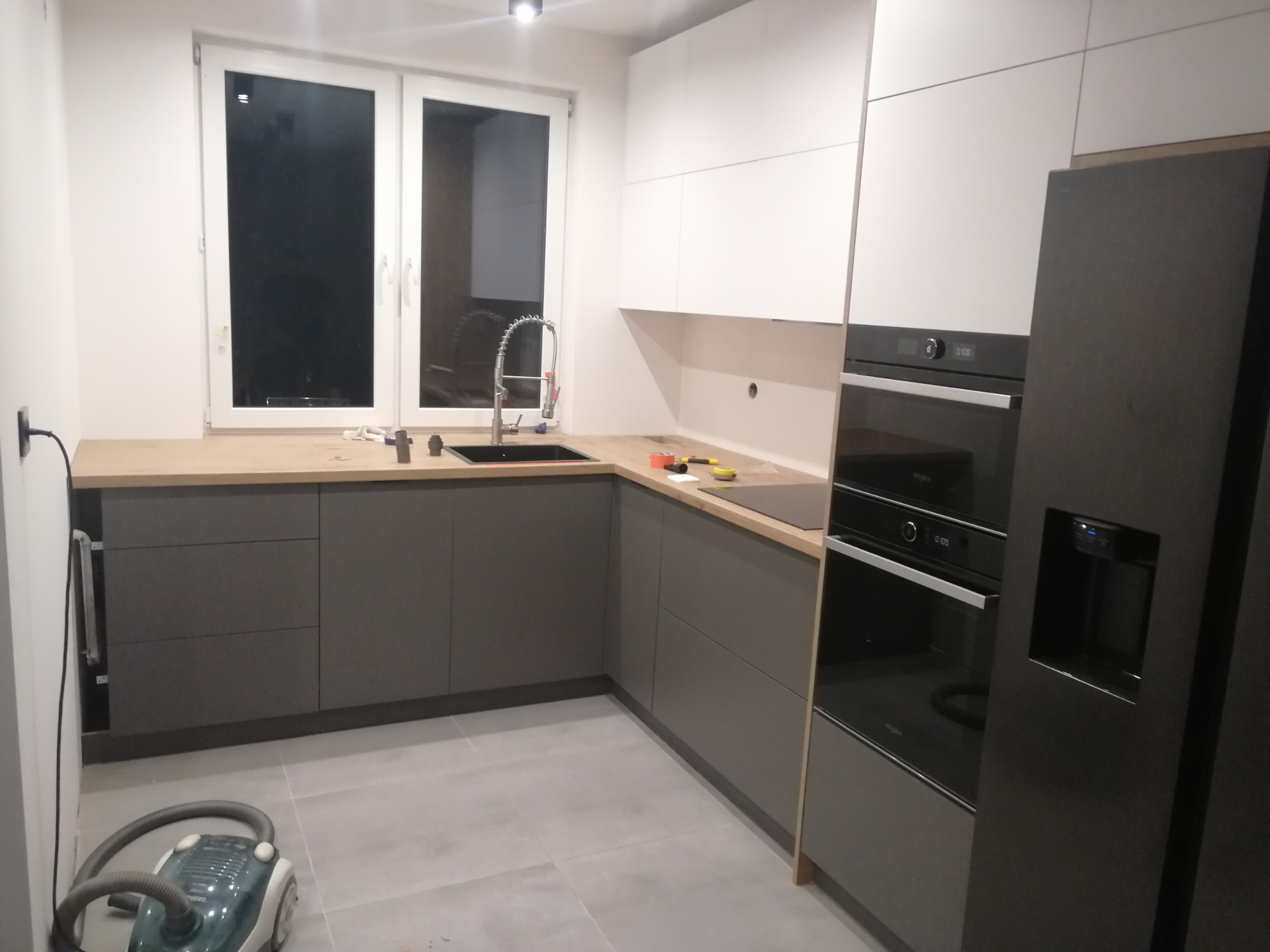 Montaż szarej, narożnej zabudowy kuchennej z białymi szafkami wiszącymi, drewnianym blatem i czarnym zlewem, widoczne narzędzia i odkurzacz.