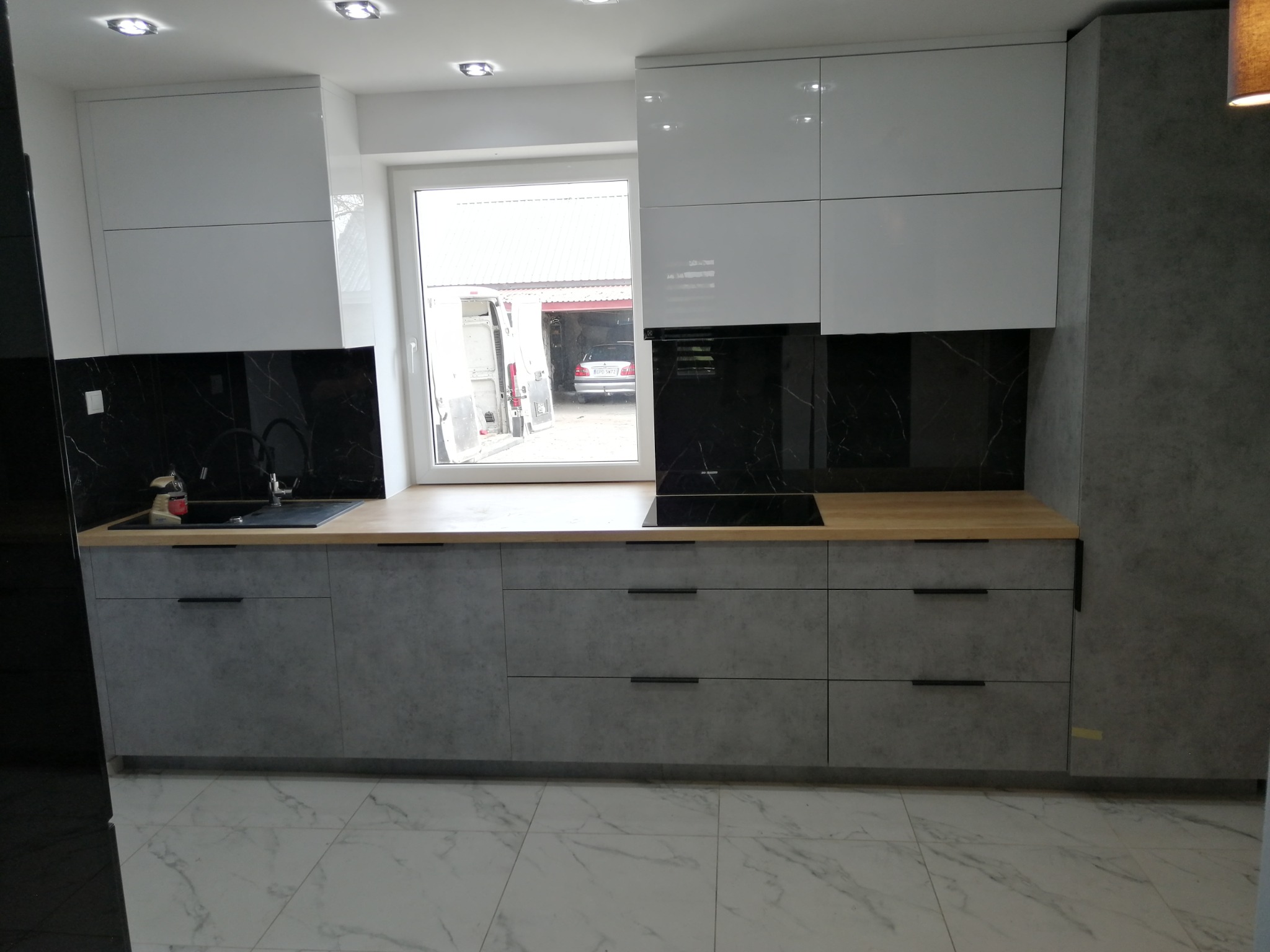 Nowoczesna kuchnia z białymi szafkami górnymi i szarymi dolnymi, drewniany blat, czarny backsplash, widok z okna na podwórko z samochodem dostawczym.