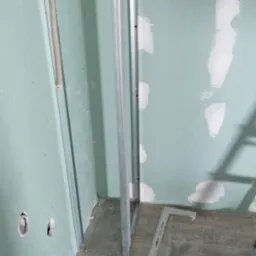 Stelaż z profili metalowych do zabudowy ściany z widoczną izolacją z wełny mineralnej, częściowo zagruntowana ściana i płytki podłogowe imitujące drewno, w tle łatka gipsowa.