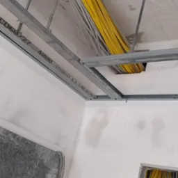 Sufit w trakcie remontu z widocznymi metalowymi profilami, żółtymi i szarymi kablami elektrycznymi oraz fragmentem ściany z widocznymi śladami po gładzi.