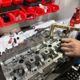 Głowica silnika z widocznymi sprężynami zaworowymi, obok narzędzia do ich demontażu w rękach mechanika, na warsztatowym stole, z czerwonymi pojemnikami na narzędzia w tle.