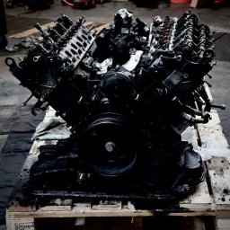 BERBER CARS SPÓŁKA Z OGRANICZONĄ ODPOWIEDZIALNOŚCIĄ - Czarny silnik samochodowy V8 spoczywający na drewnianej palecie w warsztacie, widoczne ślady oleju i smaru.