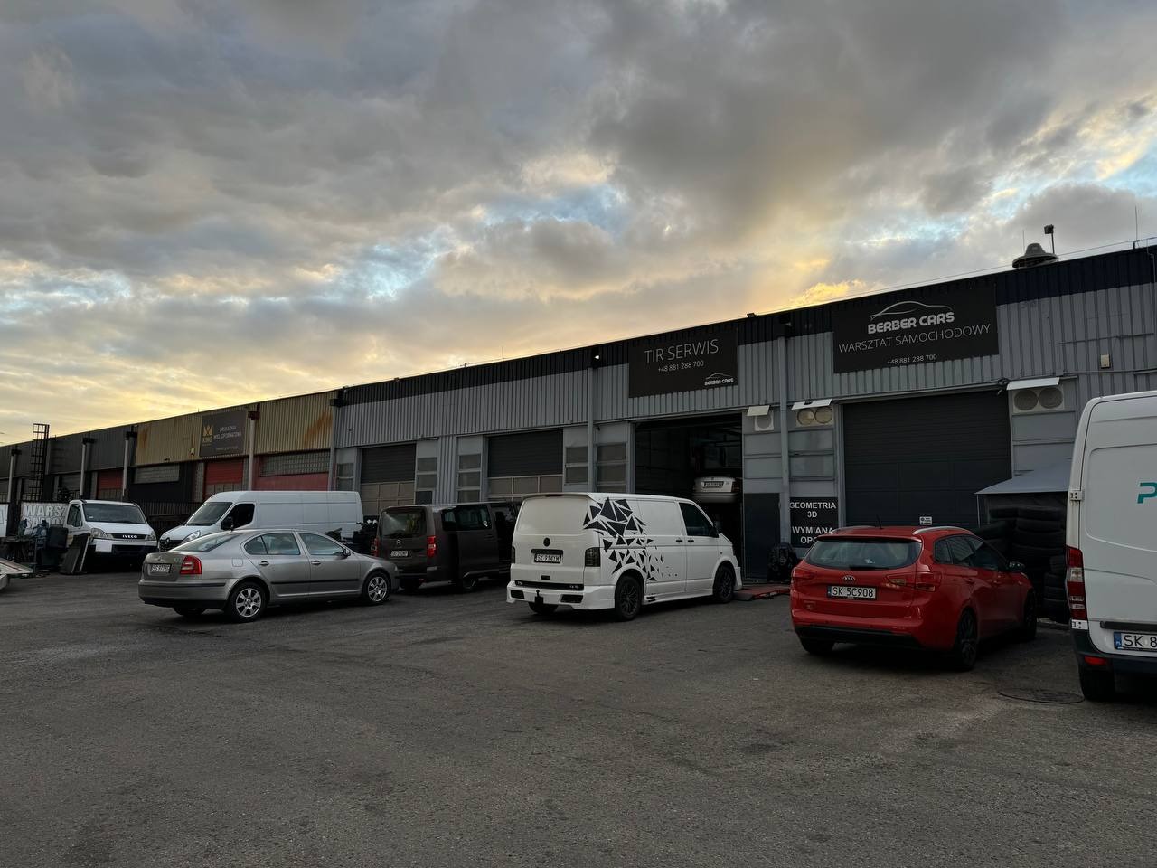Widok na warsztat samochodowy Berber Cars z zaparkowanymi pojazdami, w tym biały van z czarnym wzorem geometrycznym, czerwony hatchback i srebrna Skoda, pod pochmurnym niebem o zmierzchu.
