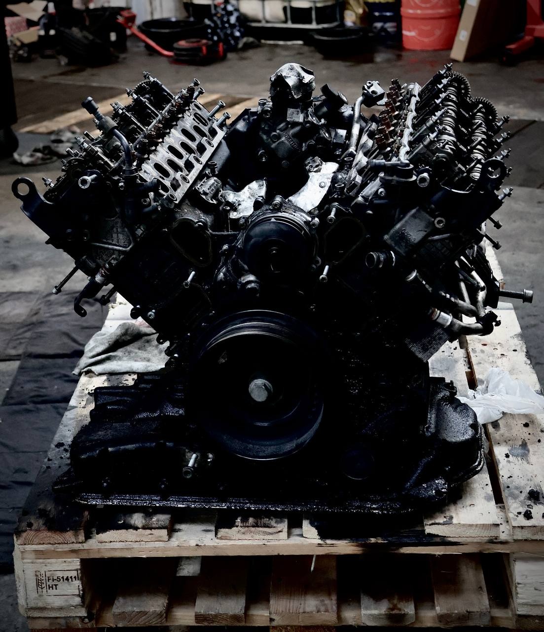 Czarny silnik samochodowy V8 spoczywający na drewnianej palecie w warsztacie, widoczne ślady oleju i smaru.
