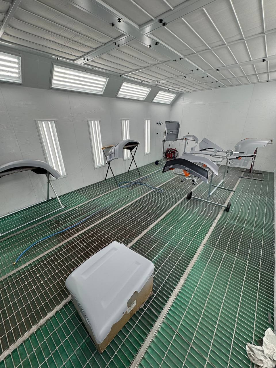 Wnętrze lakierni samochodowej z elementami karoserii przygotowanymi do lakierowania. Widoczna kratownica podłogowa i oświetlenie sufitowe.