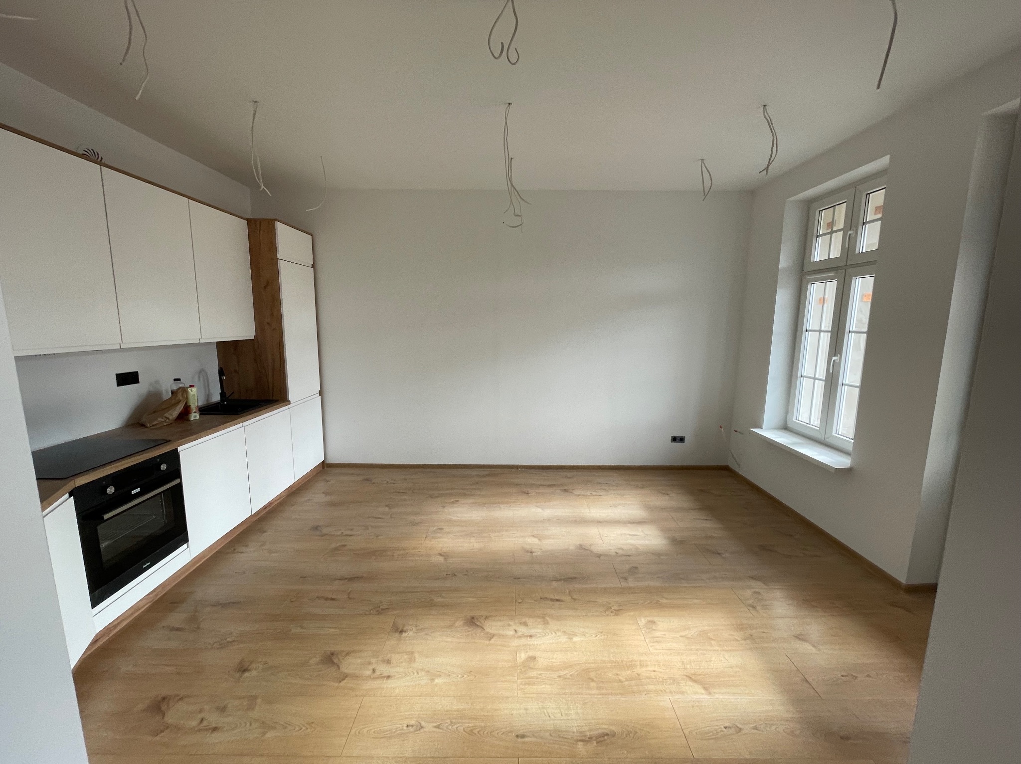 Wnętrze kuchni w trakcie prac wykończeniowych: zamontowane białe szafki z drewnianym akcentem, płyta indukcyjna i piekarnik, drewnopodobne panele na podłodze, kable zwisające z sufitu, białe ściany...