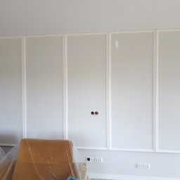 Marcin Ślęzak - Ściana z panelami dekoracyjnymi w kolorze białym, z widocznymi gniazdkami elektrycznymi i otworami na kable, zabezpieczone meble folią ochronną.