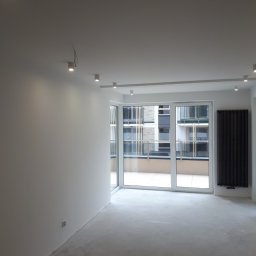 Marcin Ślęzak - Wnętrze pokoju w Kielcach przed wykończeniem: surowe ściany przygotowane pod malowanie, widoczne punkty oświetleniowe na suficie i grzejnik panelowy obok dużego okna balkonowego.