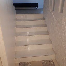 Marcin Ślęzak - Wykończone schody z białymi marmurowymi płytkami, prowadzące do jasnego pomieszczenia z czarnym meblem, ściana po prawej stronie z fakturą kamienia.