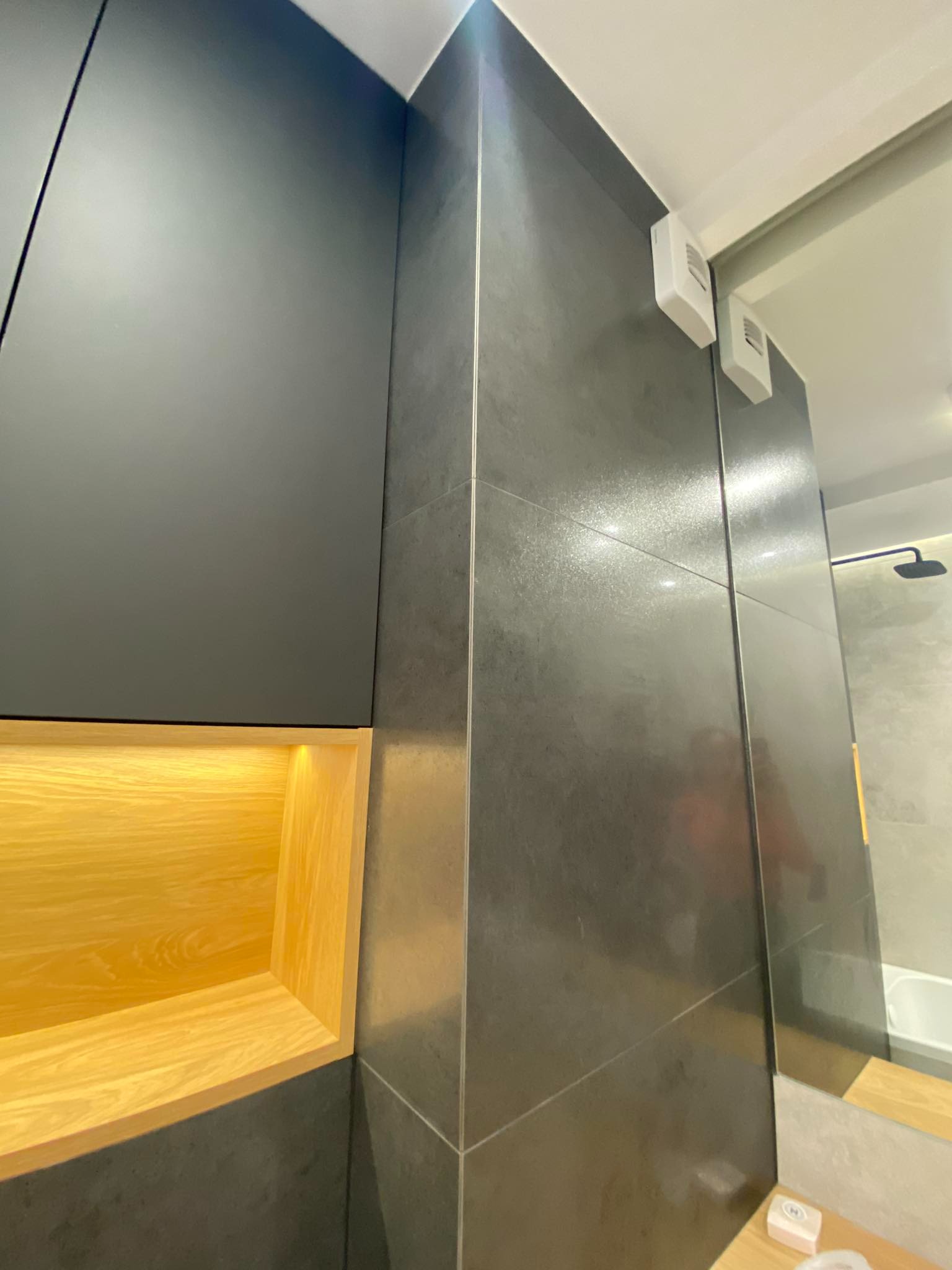 Nowoczesna łazienka z szarą, błyszczącą glazurą, zabudową meblową w kolorze grafitowym z podświetlaną, drewnianą wnęką, widoczne elementy armatury.