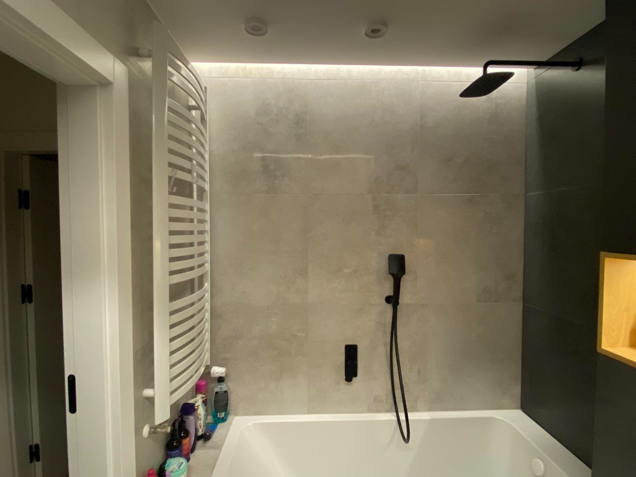 Nowoczesna łazienka z beżowymi płytkami, czarną armaturą i grzejnikiem drabinkowym, widoczne kosmetyki na wannie.