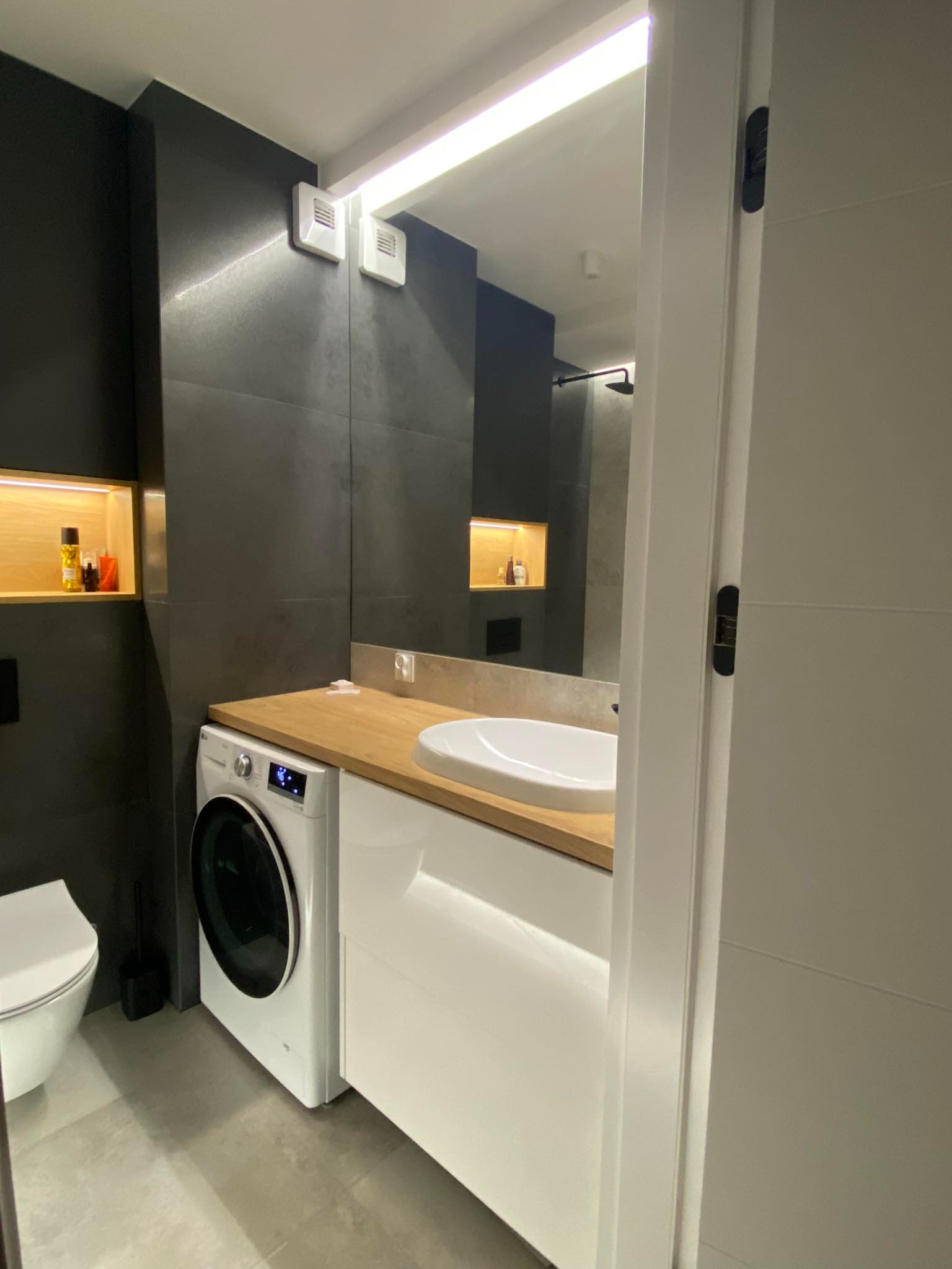 Nowoczesna łazienka z czarnymi płytkami, drewnianym blatem i pralką w zabudowie, widok zza uchylonych drzwi.