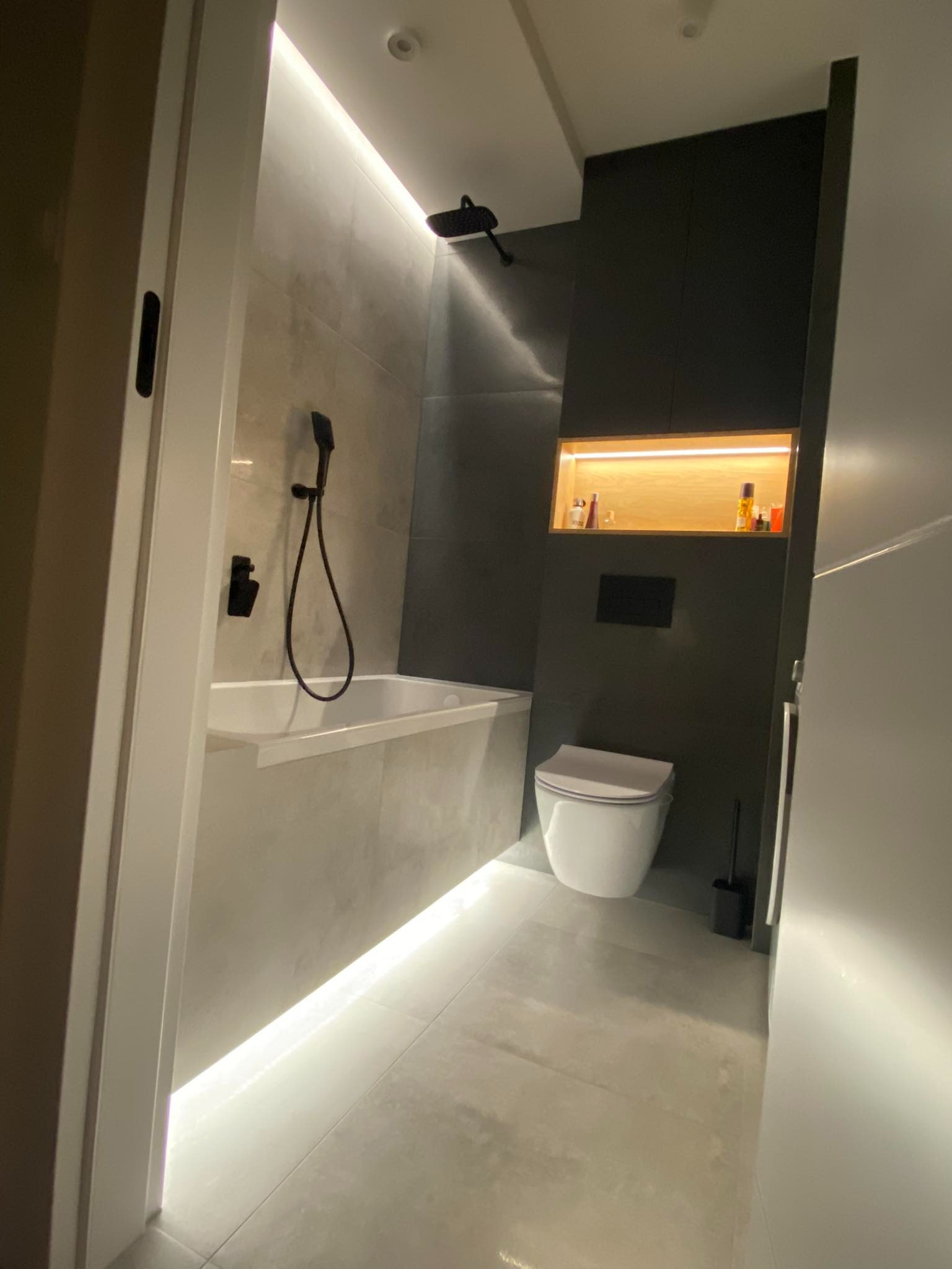 Nowoczesna łazienka z szarymi płytkami, podświetlaną wanną i wnęką, czarną armaturą i wiszącą toaletą, oświetlenie punktowe i LED.