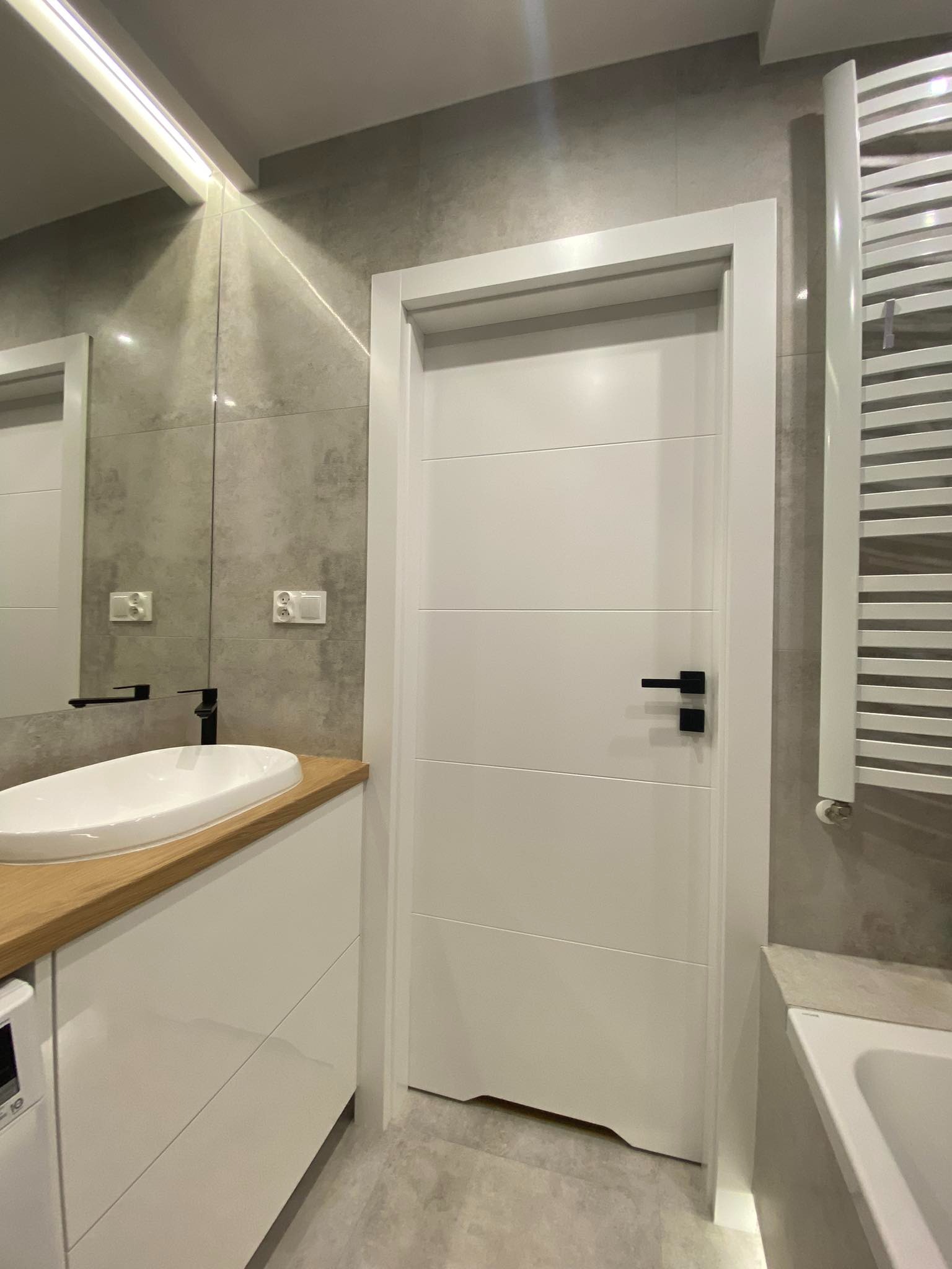 Nowoczesna łazienka z szarą płytką, białą umywalką nablatową na drewnianym blacie, białymi szafkami pod umywalką, białą ramą drzwi z czarną klamką i grzejnikiem drabinkowym.