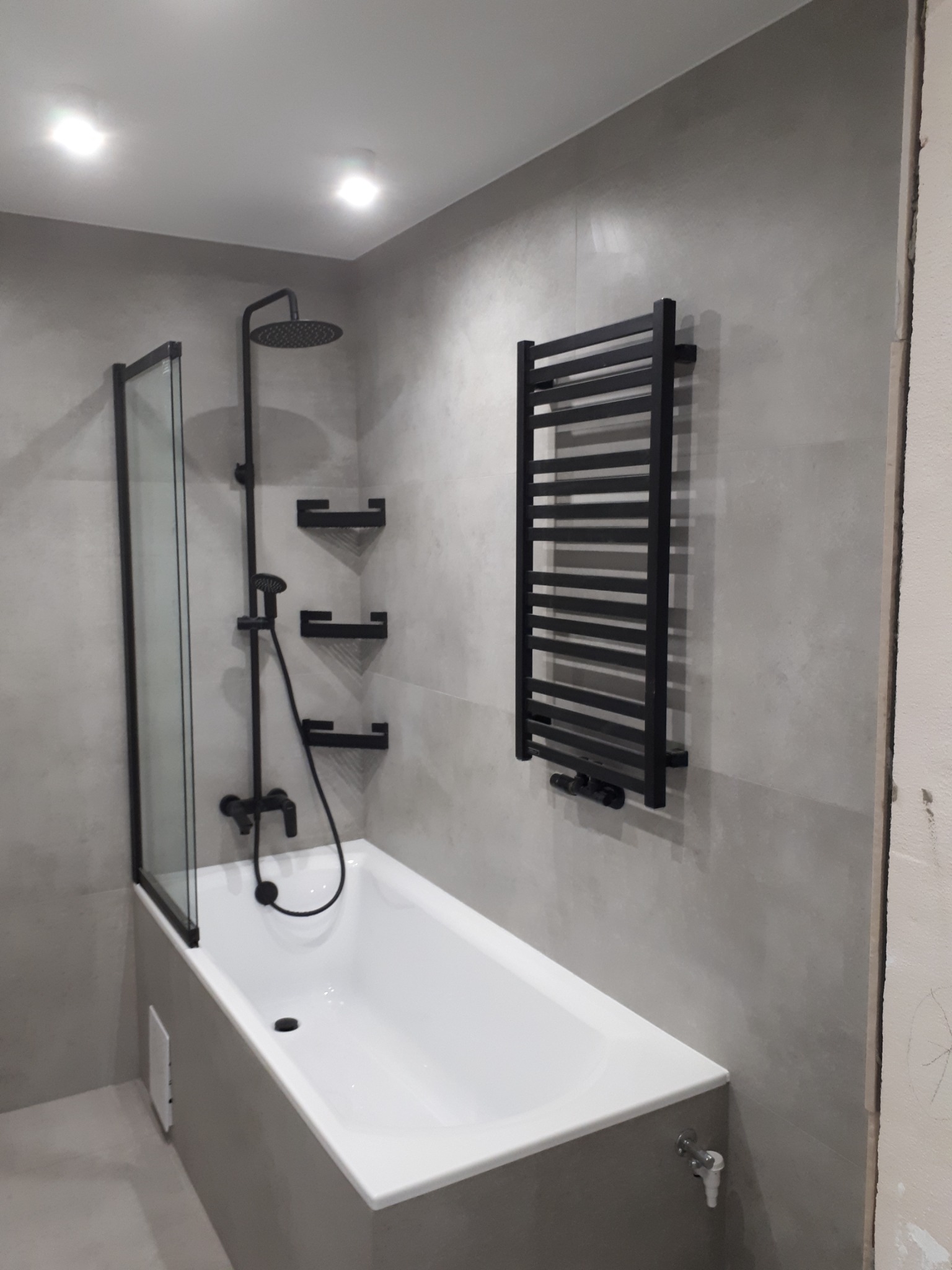 Nowoczesna łazienka z wanną, czarną armaturą i grzejnikiem drabinkowym na szarej płytce, widoczny fragment niezakończonych prac wykończeniowych.