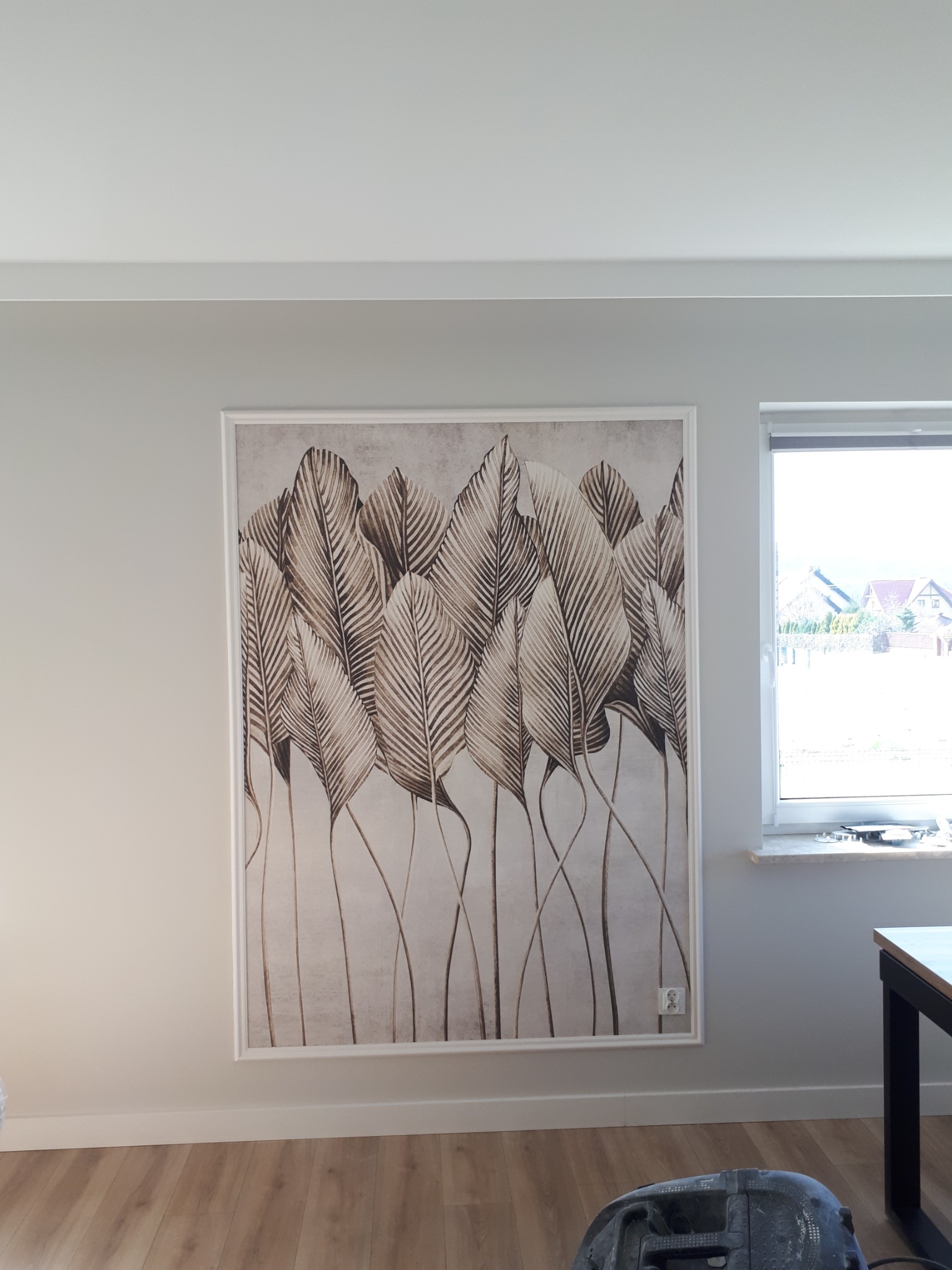 Obraz w białej ramie z motywem liści palmowych w odcieniach brązu, wiszący na szarej ścianie obok okna, z widoczną częścią parkietu i odkurzacza na podłodze.