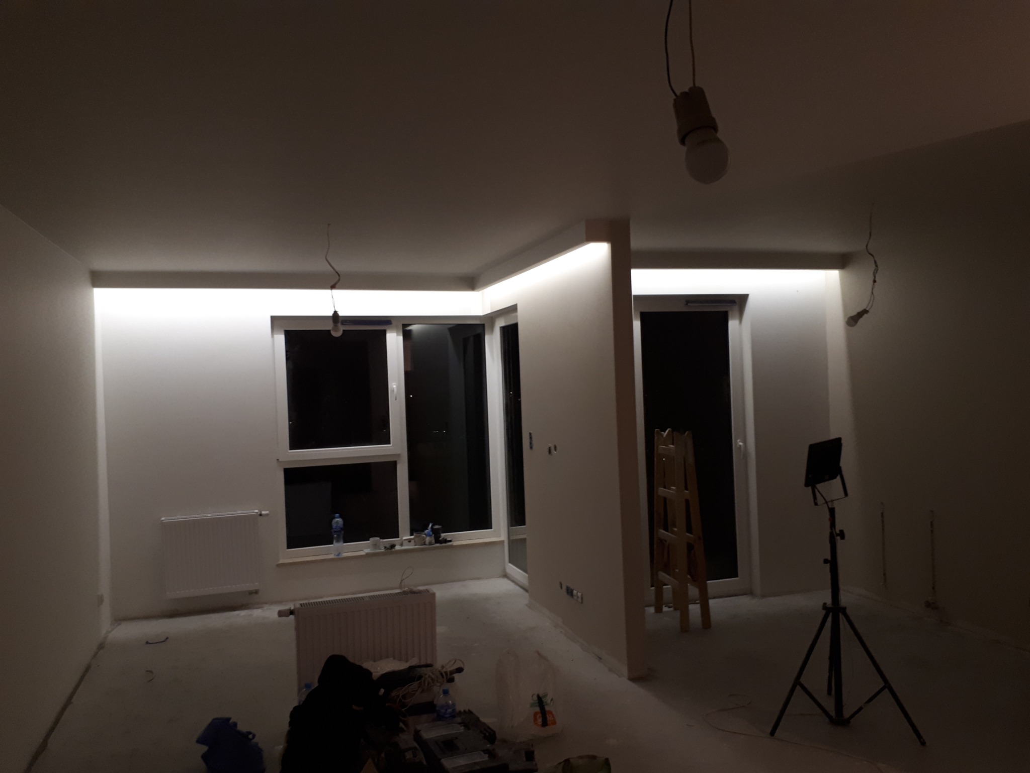 Pokój w trakcie prac wykończeniowych: białe ściany, surowy beton na podłodze, widoczne grzejniki, okna, drabina, lampa na statywie i prowizoryczne oświetlenie żarówkami na kablach. Sufit...