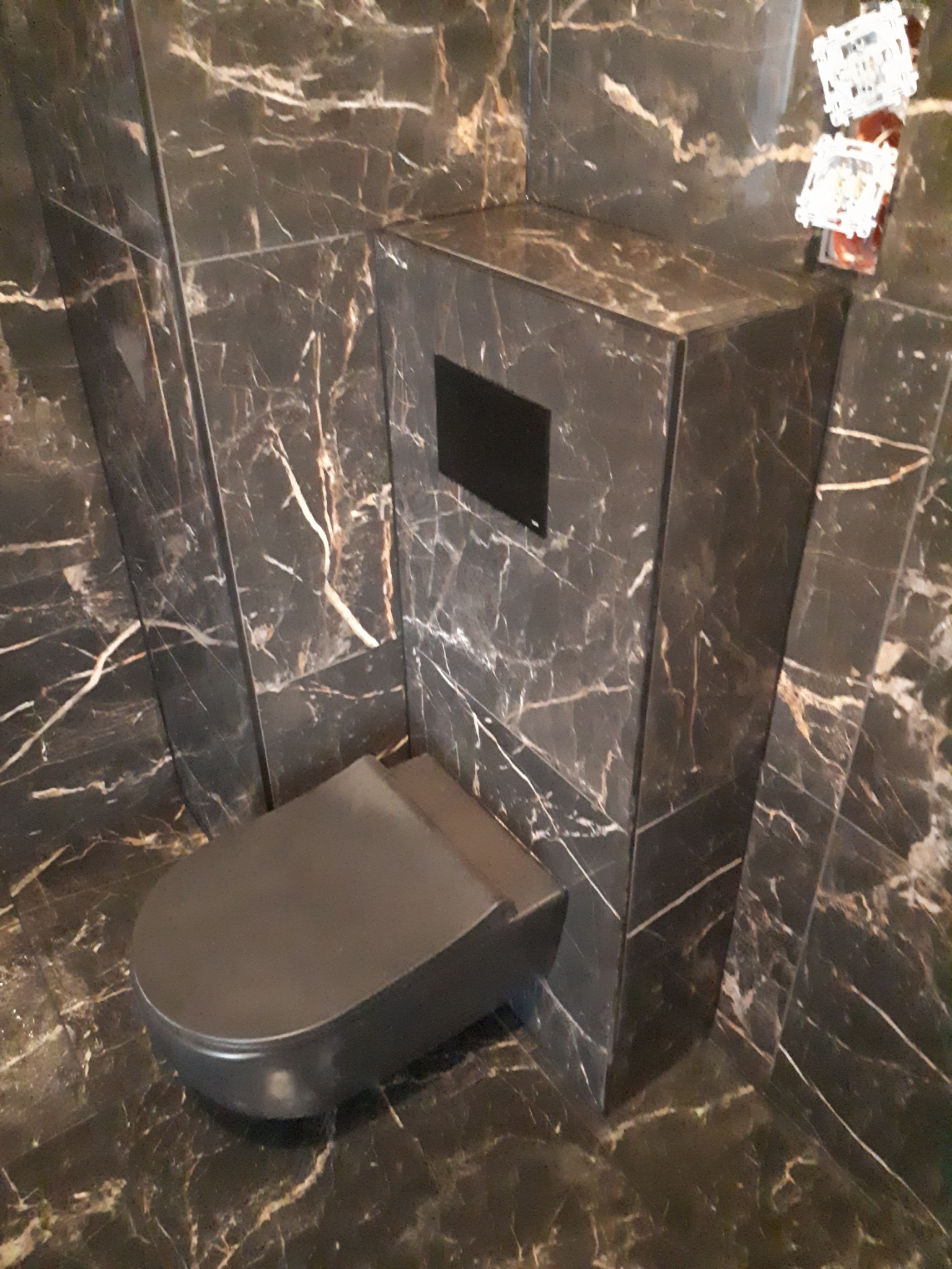 Wisząca toaleta w łazience wykończonej czarnym marmurem z białymi żyłami, z czarnym przyciskiem spłukującym i widocznymi elementami instalacji elektrycznej w rogu.