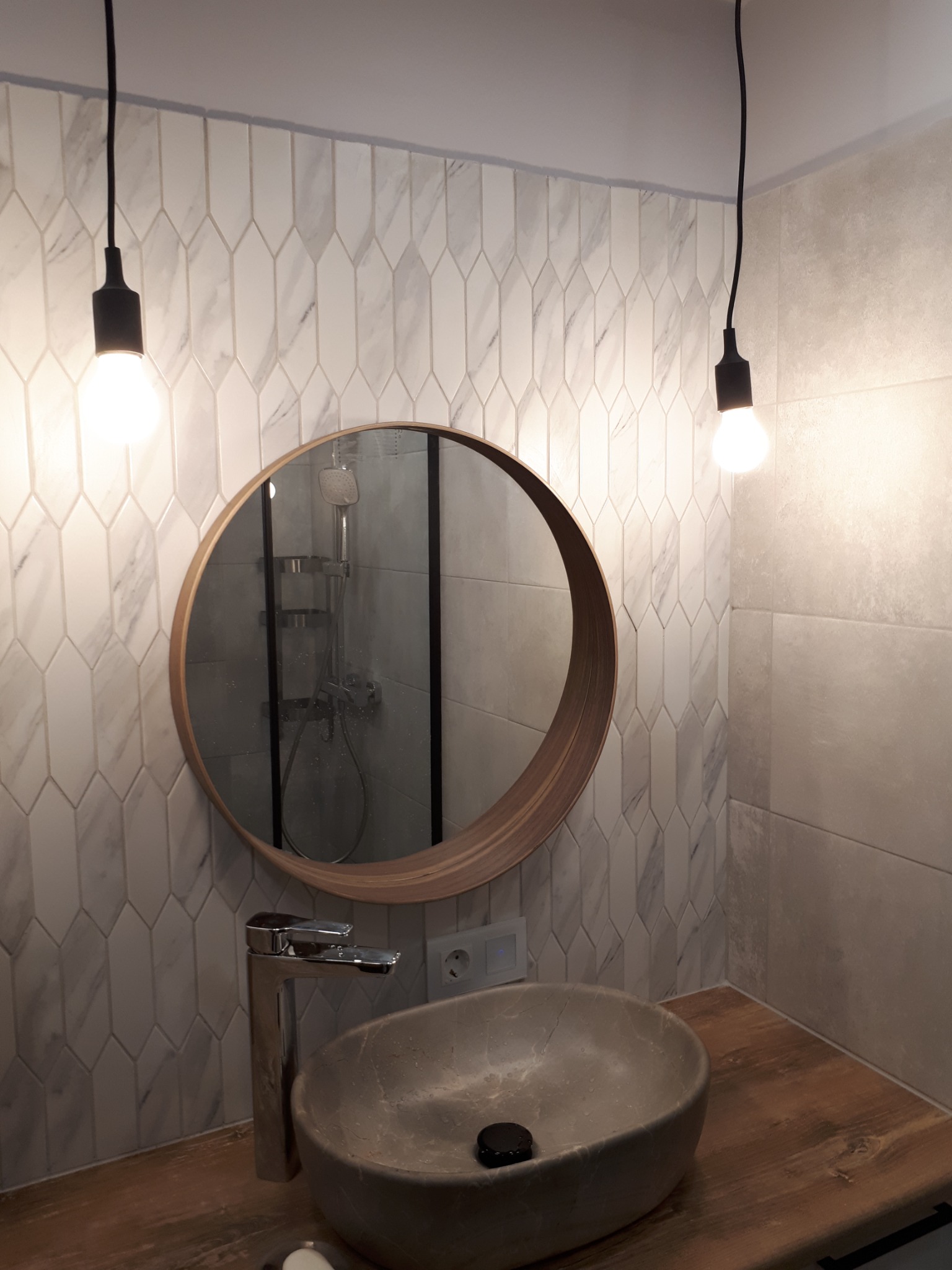 Umywalka w odcieniach szarości o nieregularnym kształcie, osadzona na drewnianym blacie, z nowoczesną baterią i okrągłym lustrem z drewnianą ramą, zawieszonym na tle ściany wyłożonej białymi...