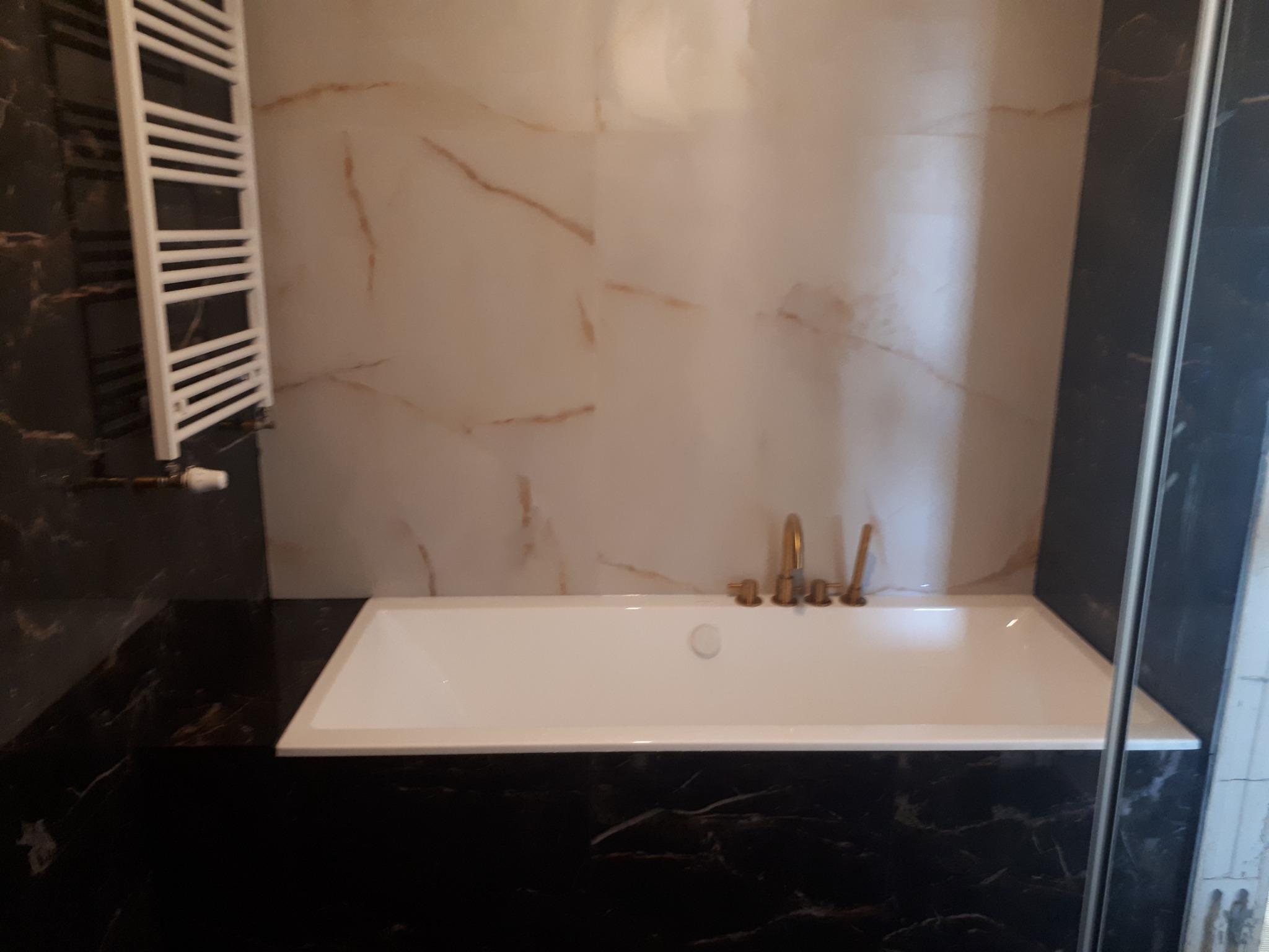 Biała wanna z złotą armaturą na tle płytek imitujących marmur, obok grzejnik drabinkowy i ciemne marmurowe wykończenie.