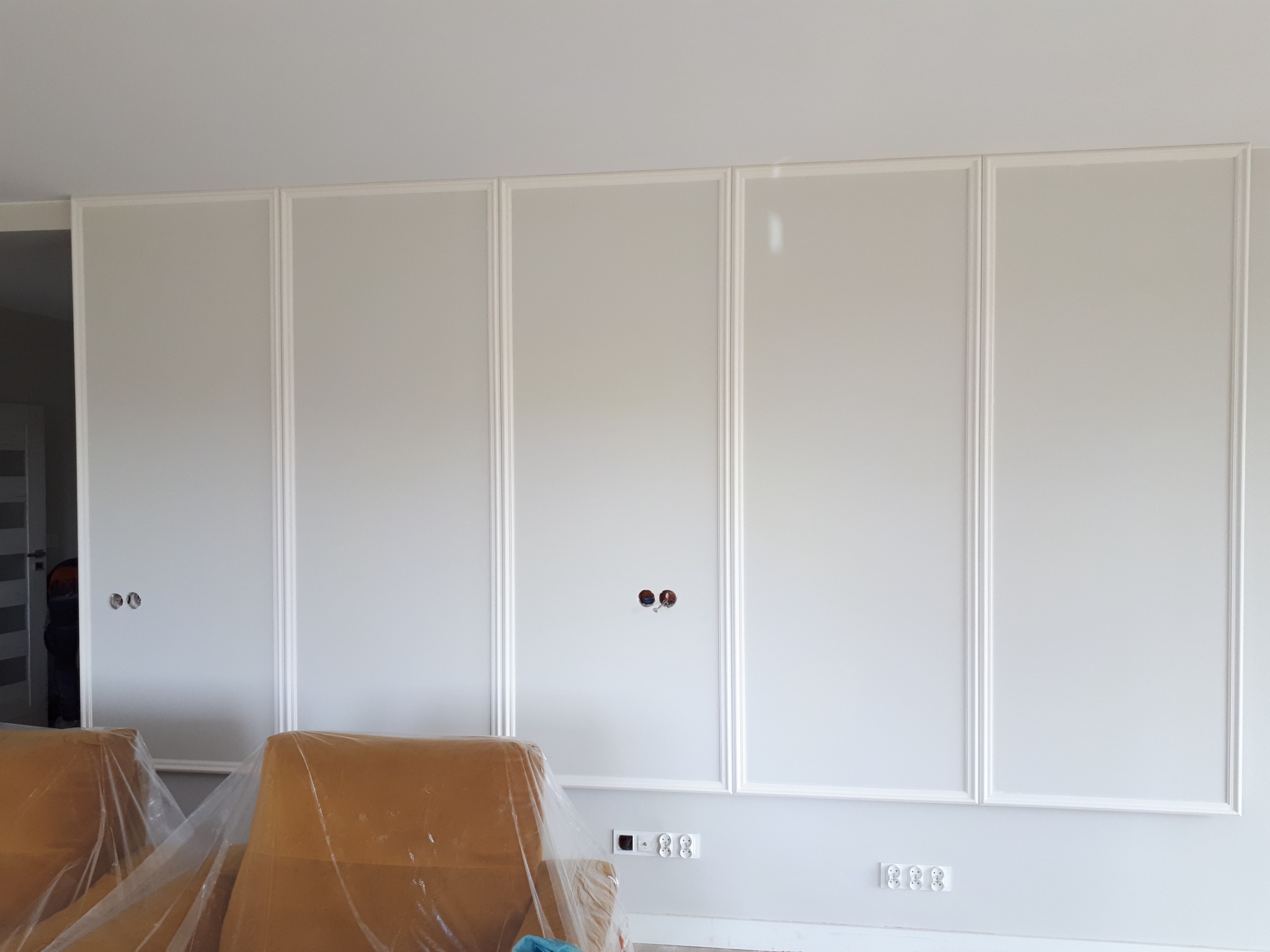 Ściana z panelami dekoracyjnymi w kolorze białym, z widocznymi gniazdkami elektrycznymi i otworami na kable, zabezpieczone meble folią ochronną.