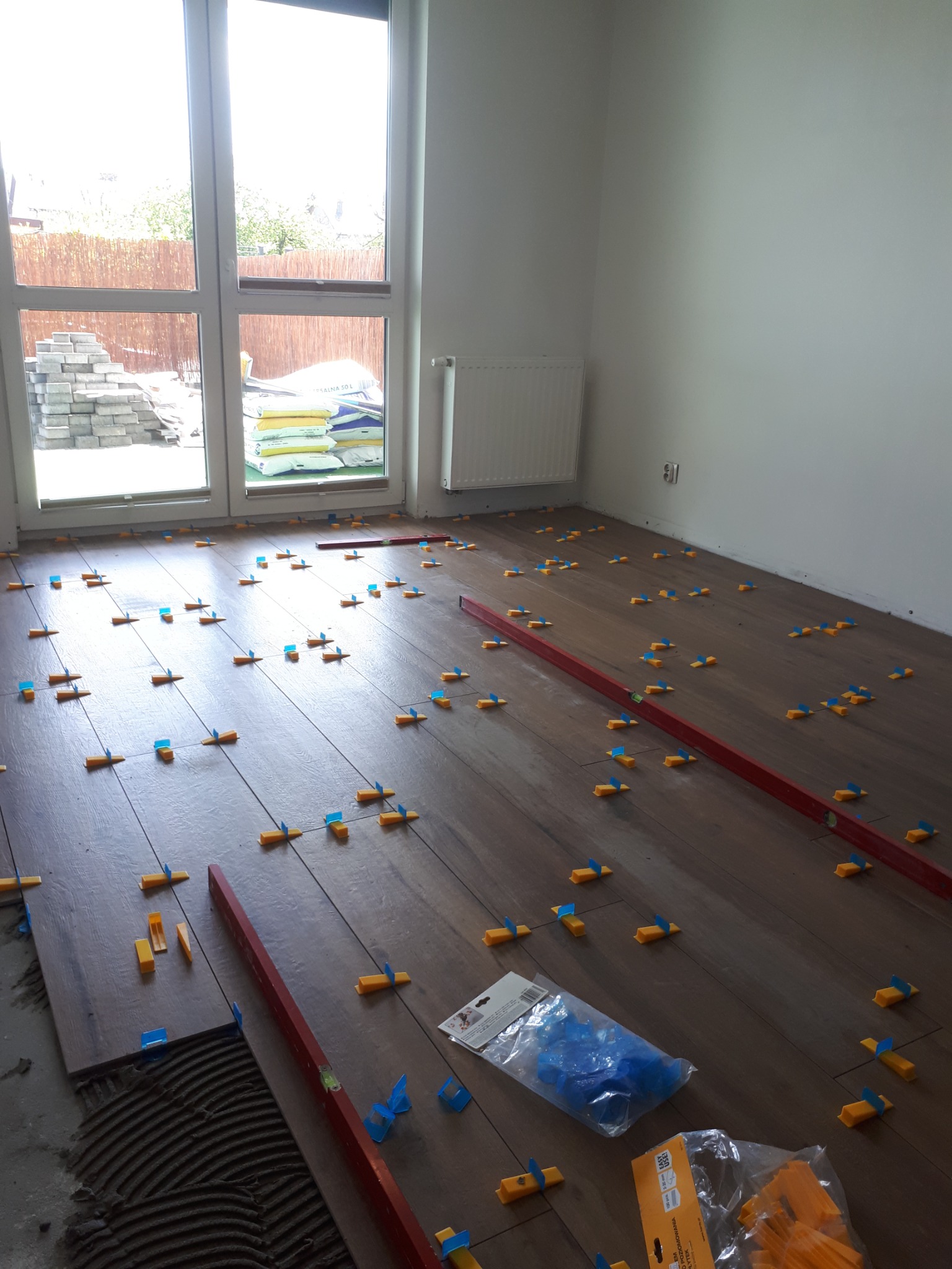 Układanie ciemnych paneli podłogowych z użyciem poziomicy i systemu poziomowania płytek; w tle okno z widokiem na materiały budowlane.