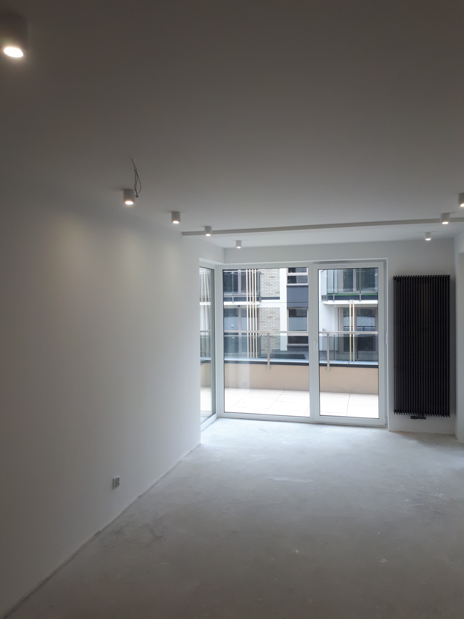 Wnętrze pokoju w Kielcach przed wykończeniem: surowe ściany przygotowane pod malowanie, widoczne punkty oświetleniowe na suficie i grzejnik panelowy obok dużego okna balkonowego.