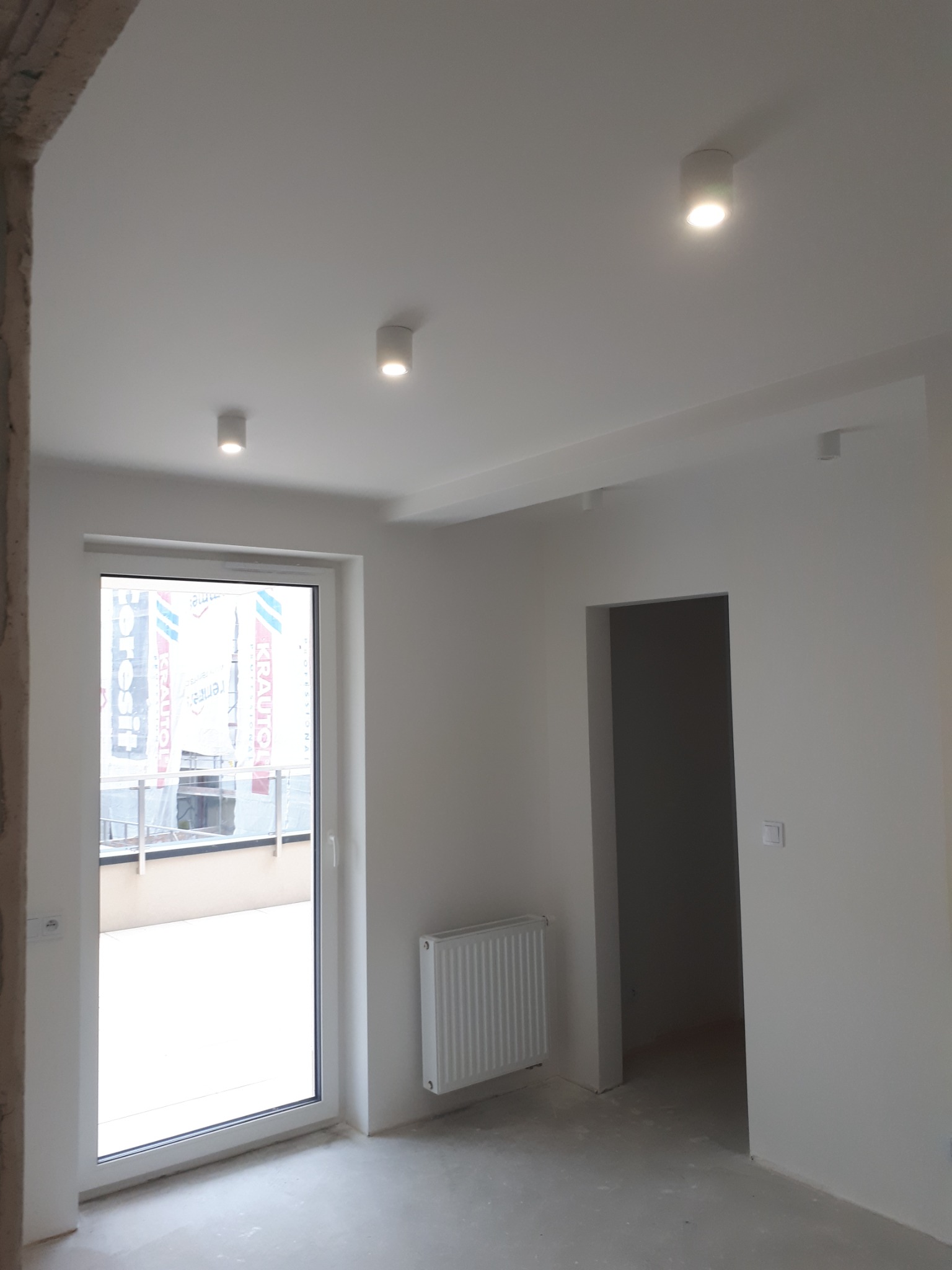 Wnętrze w trakcie prac wykończeniowych: białe ściany, surowa podłoga, grzejnik, okno balkonowe z widokiem na balkon i trzy punktowe lampy sufitowe.