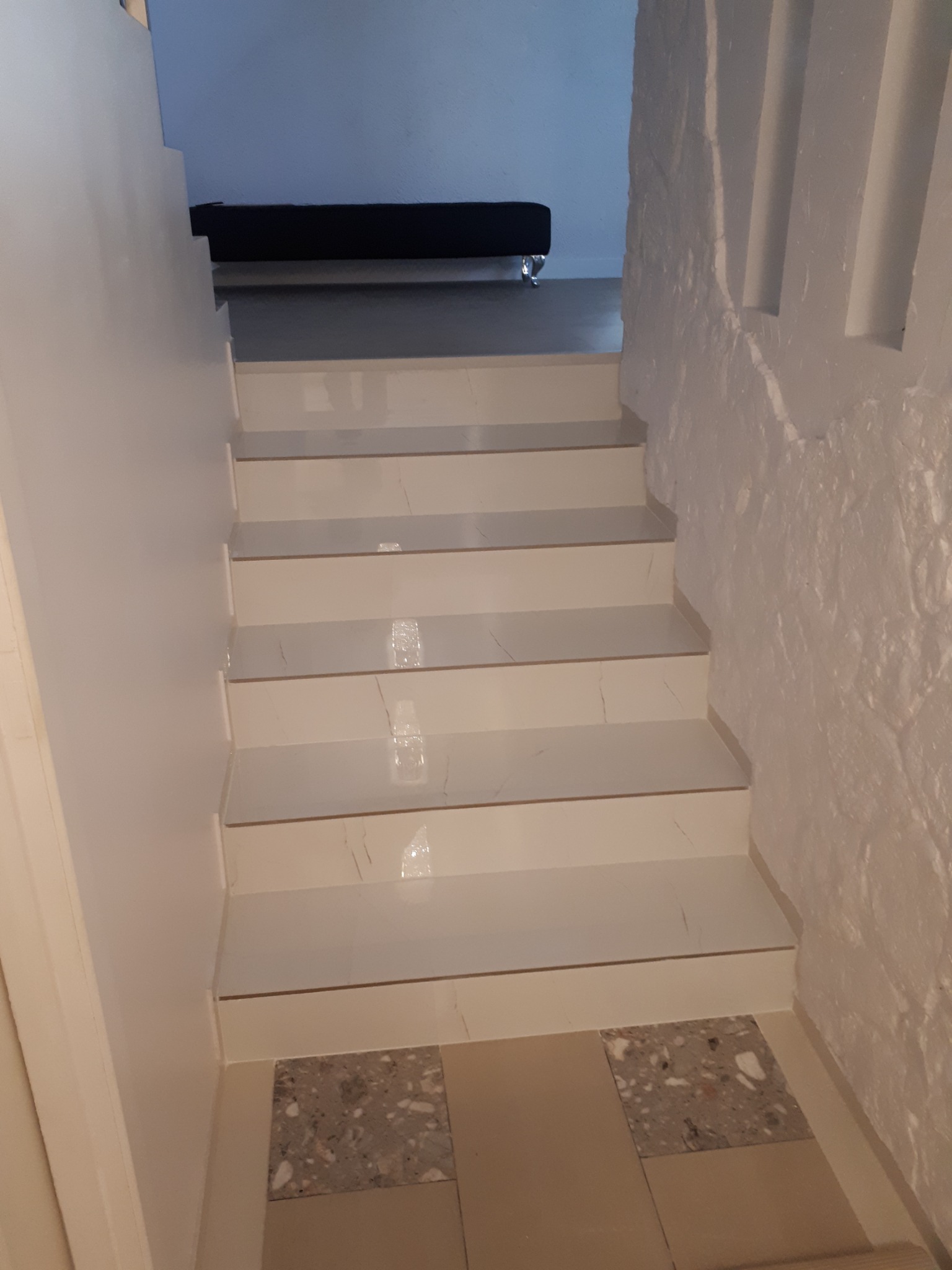 Wykończone schody z białymi marmurowymi płytkami, prowadzące do jasnego pomieszczenia z czarnym meblem, ściana po prawej stronie z fakturą kamienia.