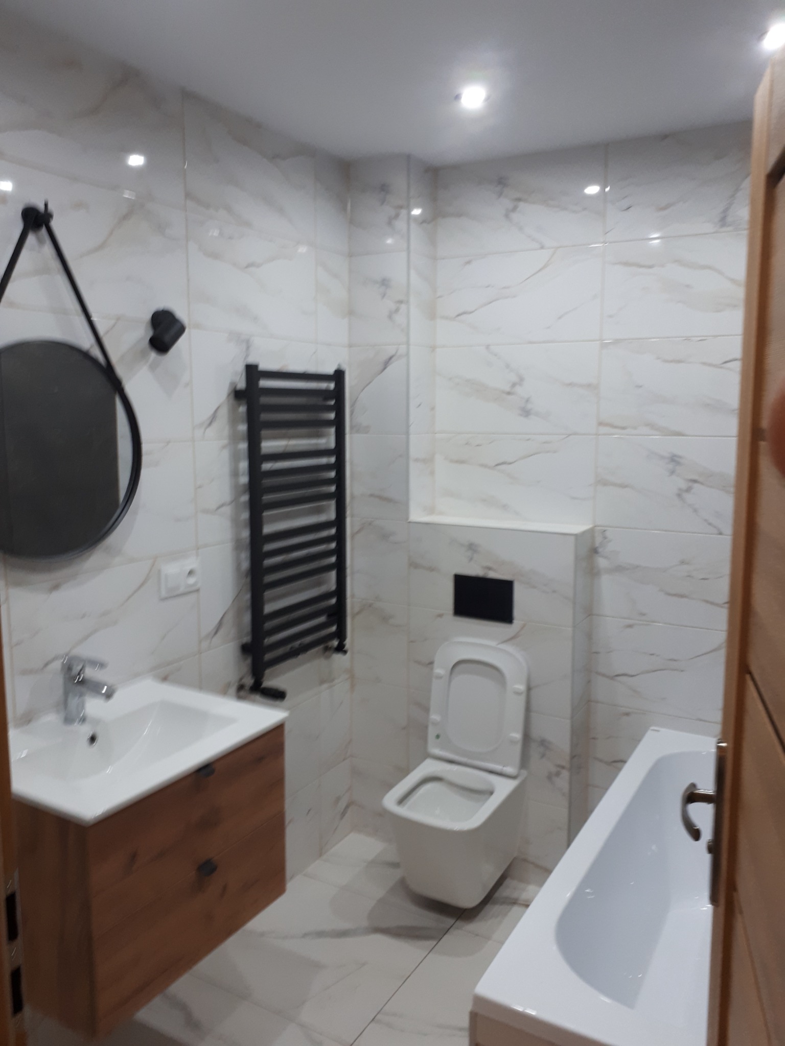 Nowoczesna łazienka z marmurowymi płytkami, wiszącą szafką z umywalką o drewnianym wykończeniu, okrągłym lustrem na czarnym uchwycie, grzejnikiem drabinkowym i podwieszaną toaletą.