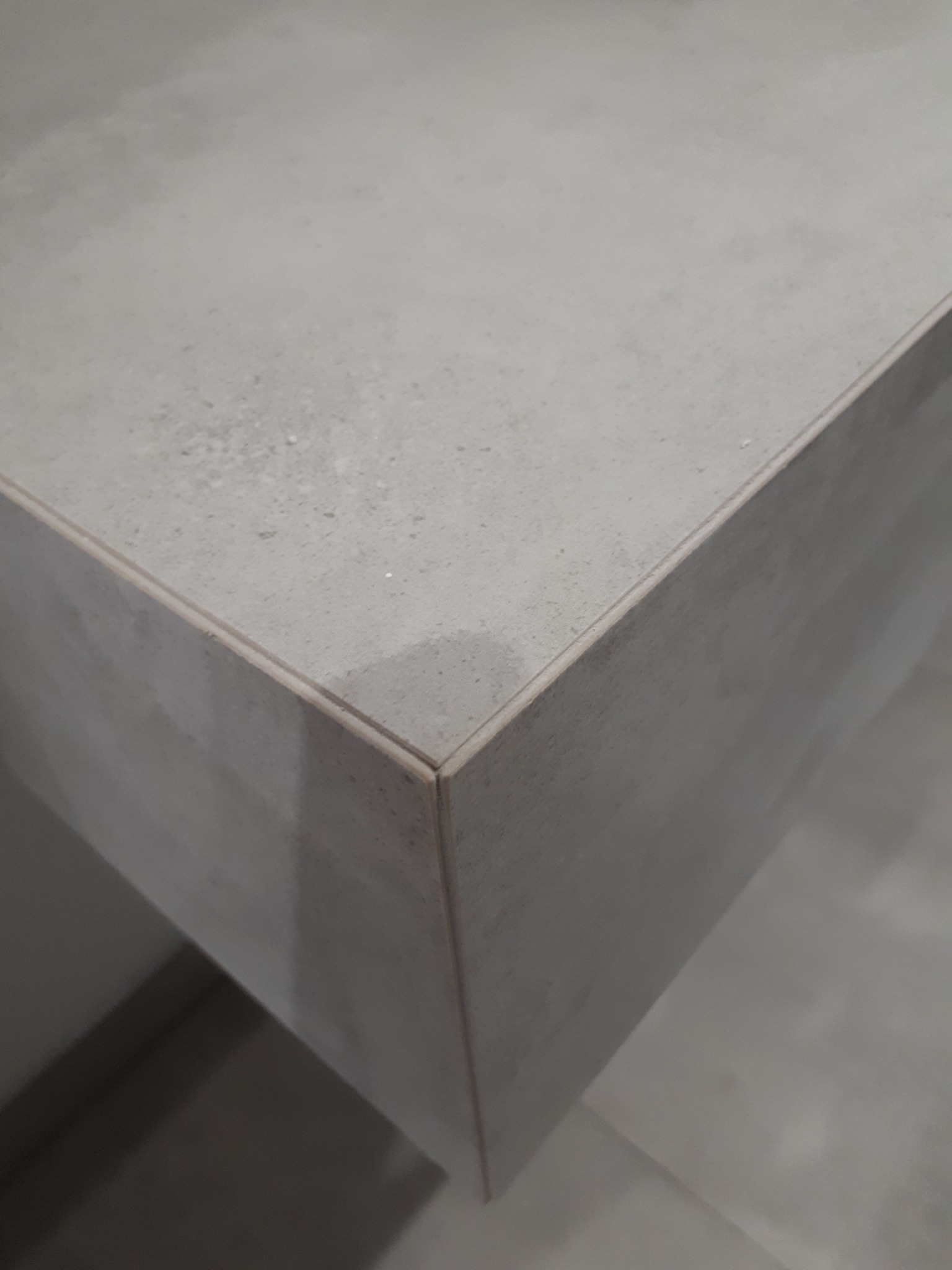 Szczegółowe ujęcie rogu blatu z szarego, matowego kamienia kompozytowego, wykończonego delikatną, jasną listwą. Widoczna faktura materiału.
