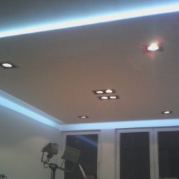 Aldonbud - Podwieszany sufit z oświetleniem LED i punktowymi lampami w nowoczesnym pokoju, widoczny statyw oświetleniowy w rogu.