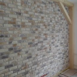 Aldonbud - Ściana wykończona dekoracyjną cegłą w odcieniach beżu, szarości i brązu, z widoczną drewnianą konstrukcją ramy. Widoczne gniazdka elektryczne w dolnej części ściany.