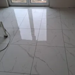 Błyszcząca, biała podłoga z płytek imitujących marmur, świeżo po ułożeniu, widoczny grzejnik elektryczny i okno z widokiem na gruzowisko.