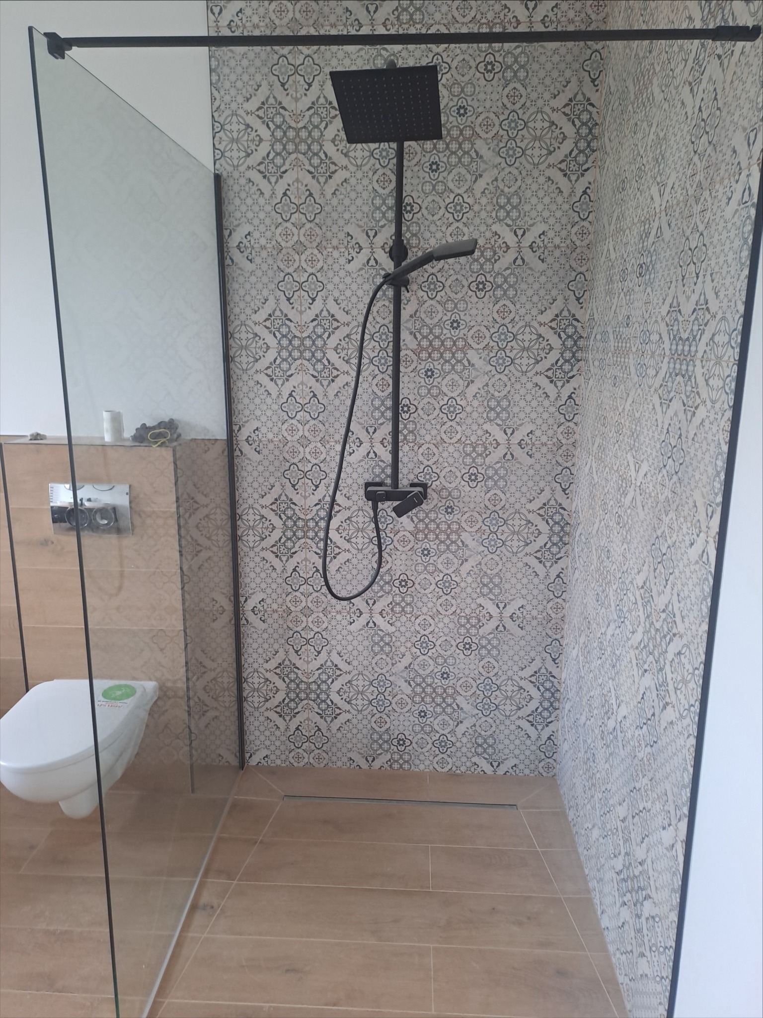 Nowoczesna kabina prysznicowa walk-in z czarną armaturą i wzorzystymi płytkami na ścianie, widoczny odpływ liniowy i toaleta podwieszana.